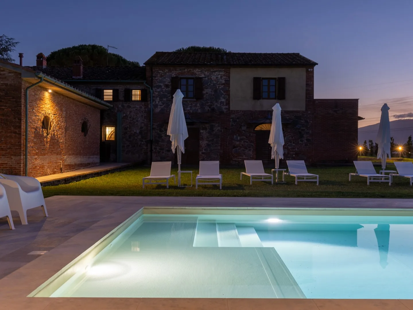 Una piscina exterior iluminada está frente a un edificio de piedra al anochecer.