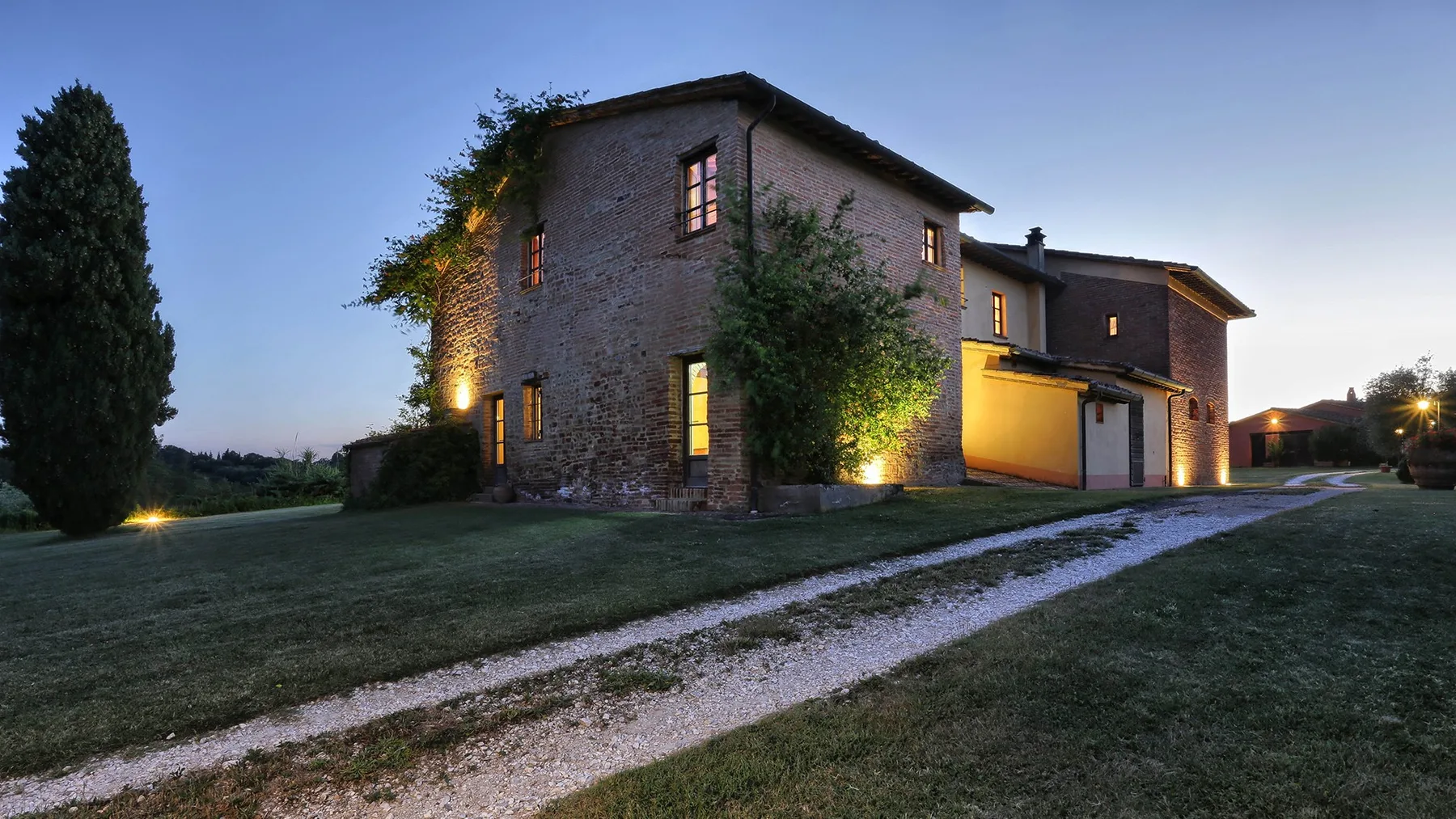 A stone house stands beside a gravel driveway on a grassy lawn at dusk.