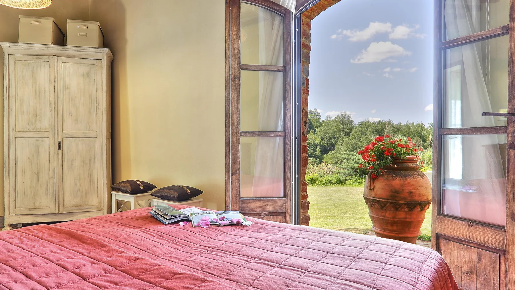 A bedroom with a pink bedspread and open doors overlooking a garden.
