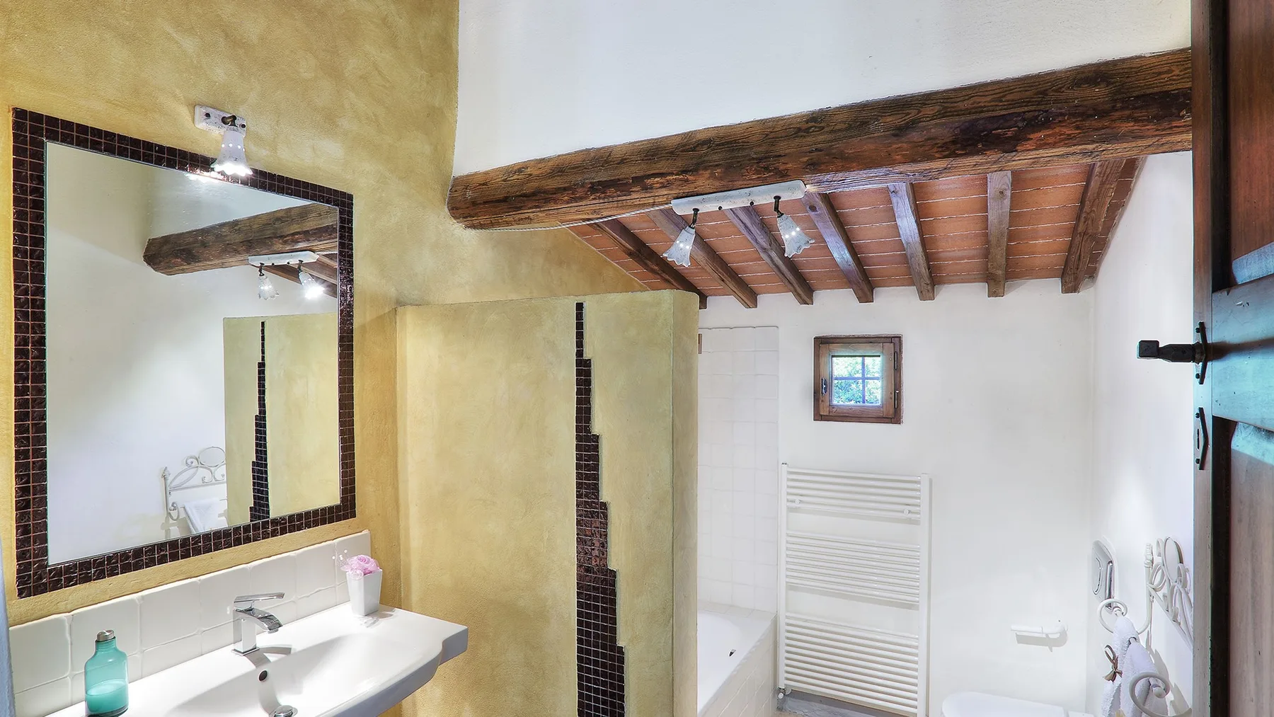 A bathroom with a sink, mirror, and shower beneath exposed wooden ceiling beams.