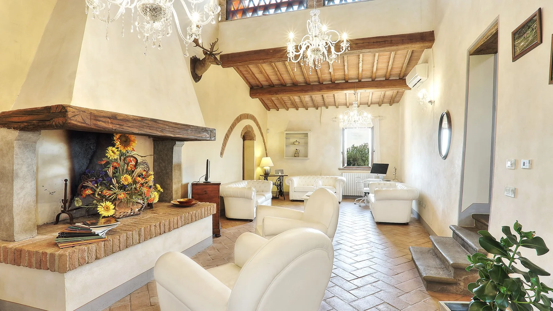 A bright living room with white seating, a large fireplace, and exposed wooden ceiling beams.