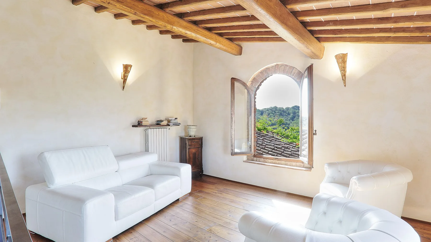 A bright living room with white seating, an arched window, and exposed wooden ceiling beams.