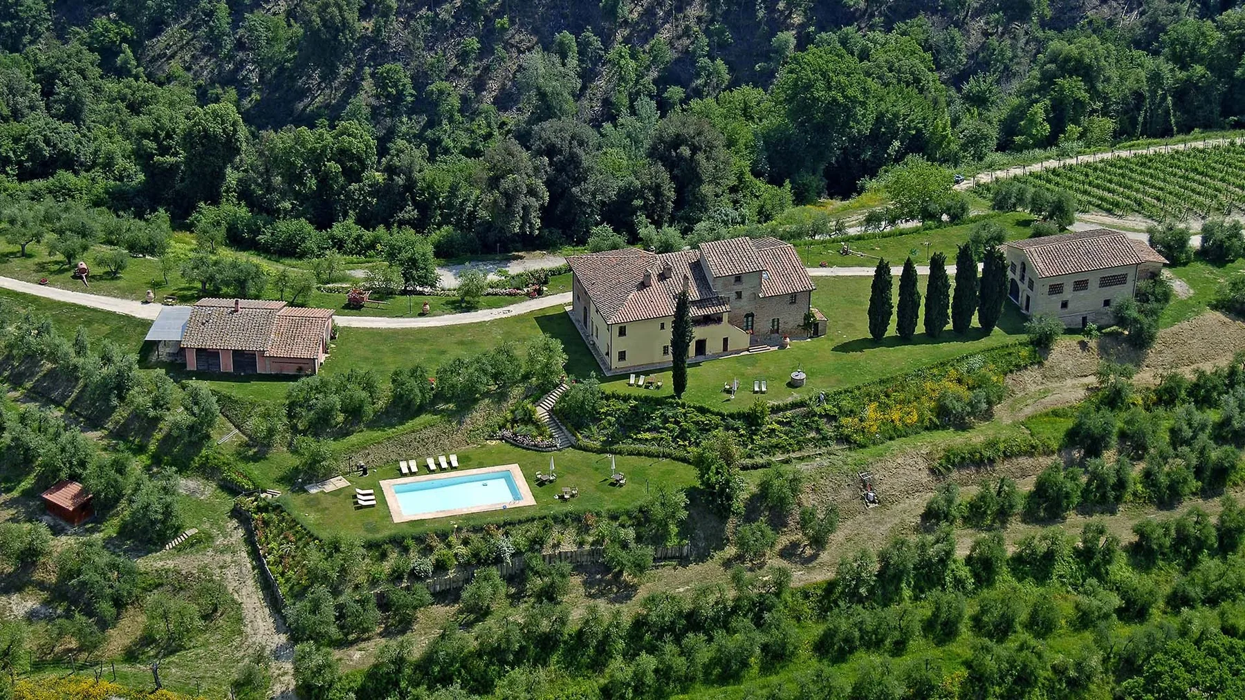 A rural property sits among wooded hills, gardens, and a swimming pool.