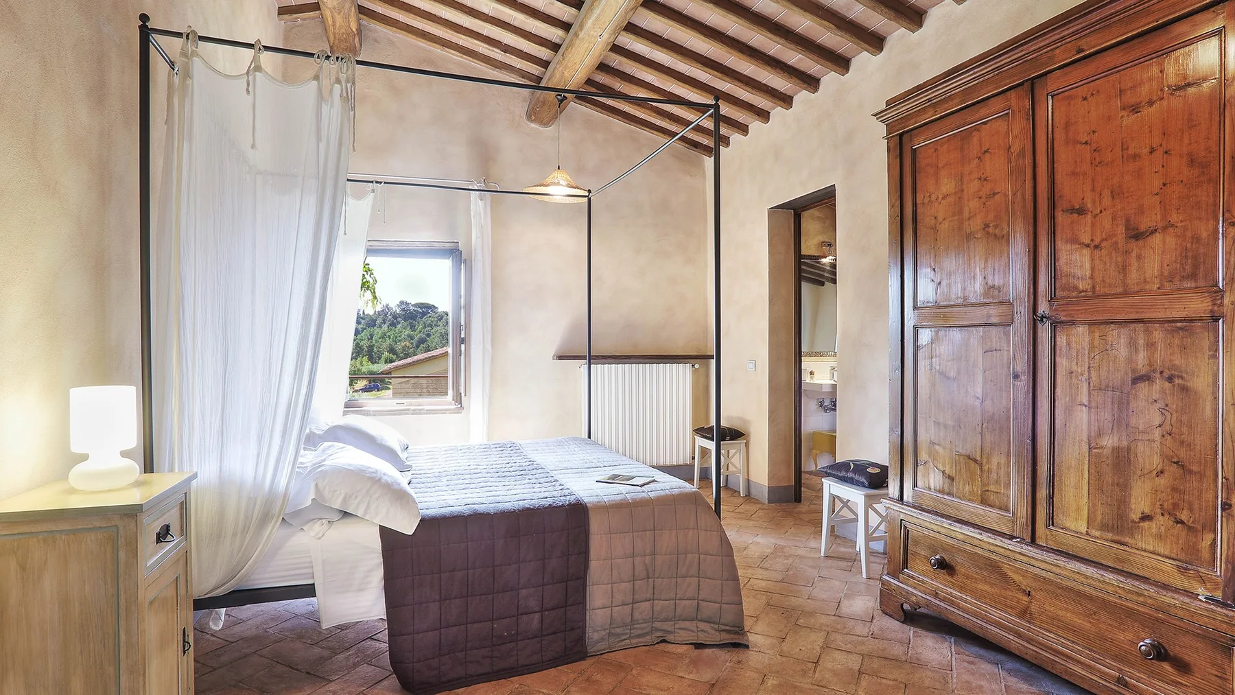 A bedroom with a canopy bed, a large wooden wardrobe, and a window.
