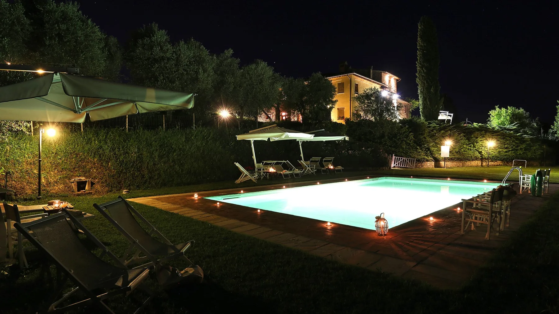 A lit outdoor swimming pool sits beside lounge seating in a garden at night.