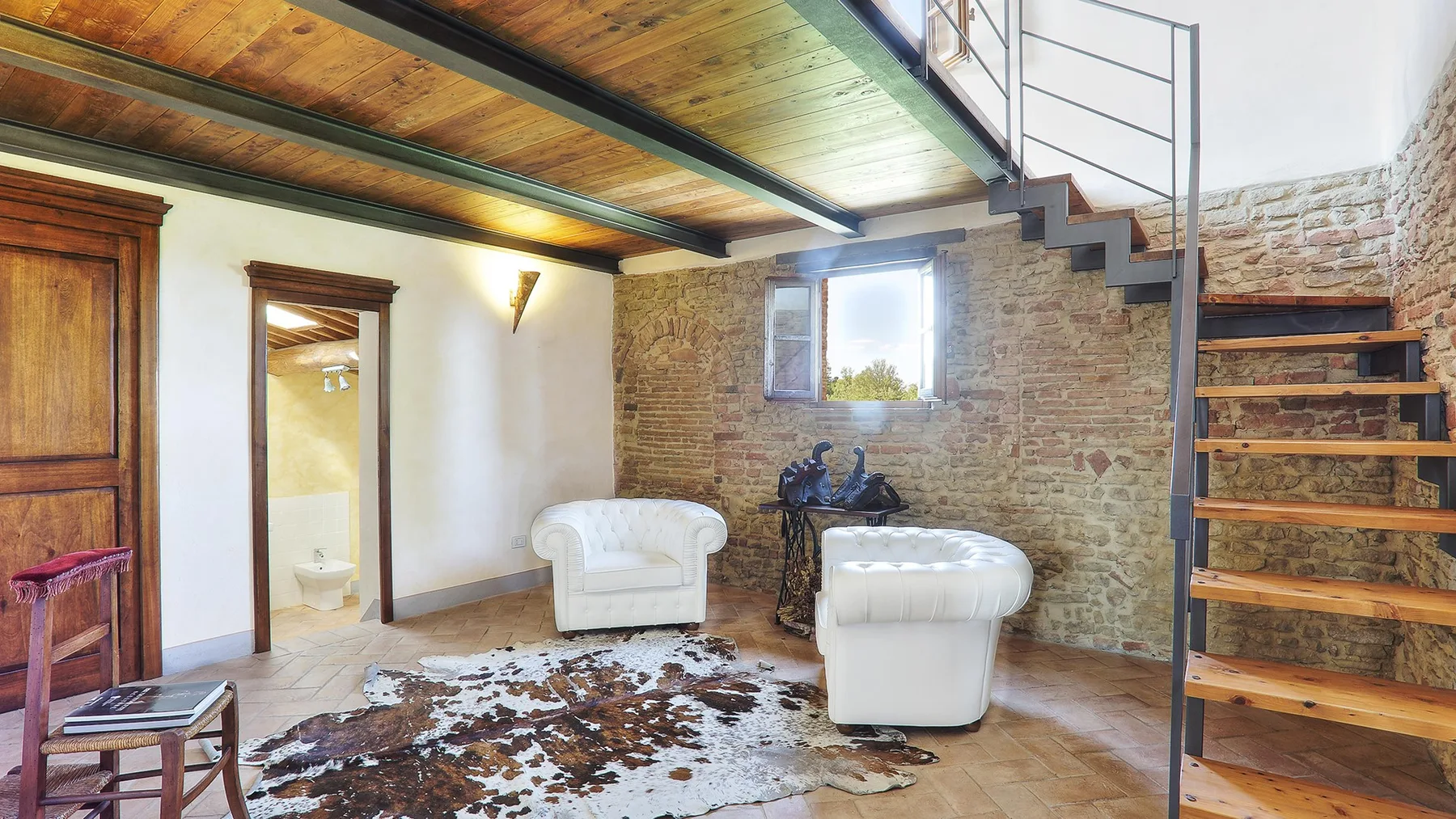 A rustic living area has white seating beneath a wooden loft and staircase.