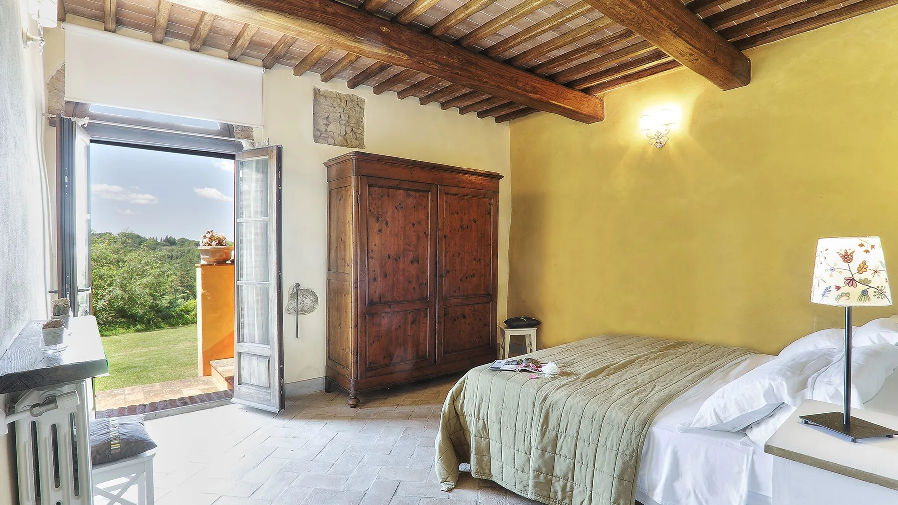 A bedroom with a wooden wardrobe, a bed, and an open doorway to greenery.