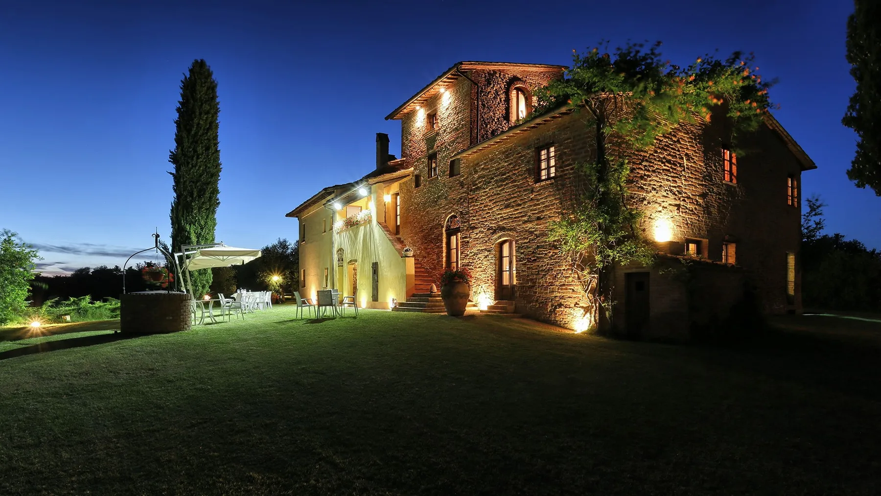 A stone building is illuminated at dusk beside a lawn and tall tree.