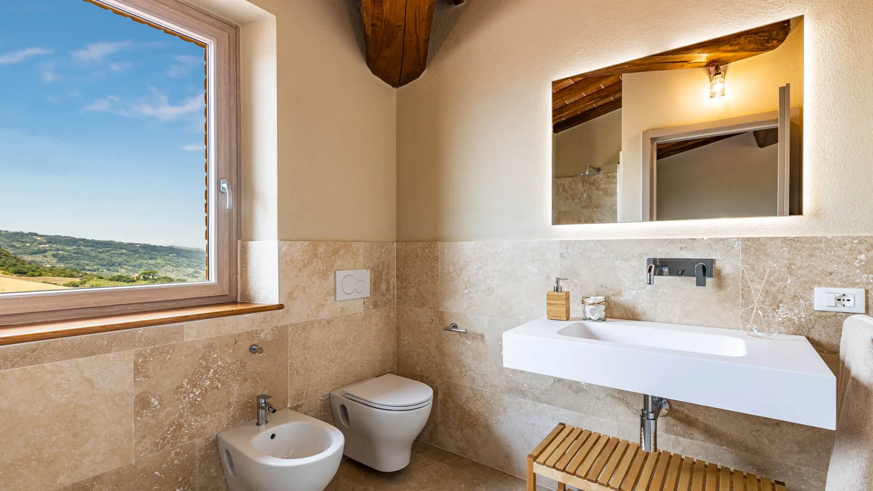 A bright bathroom with a sink, toilet, and a window overlooking the landscape.