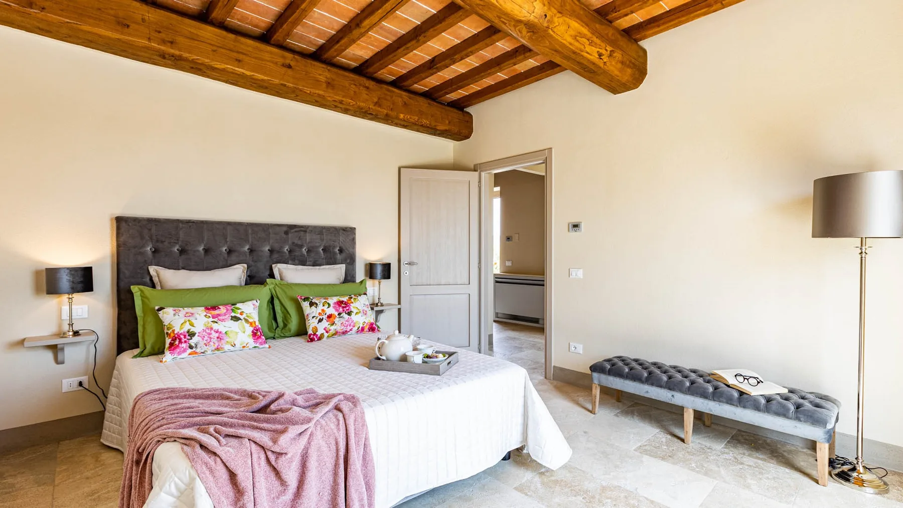A bedroom with a large bed, floral pillows, and exposed wooden ceiling beams.