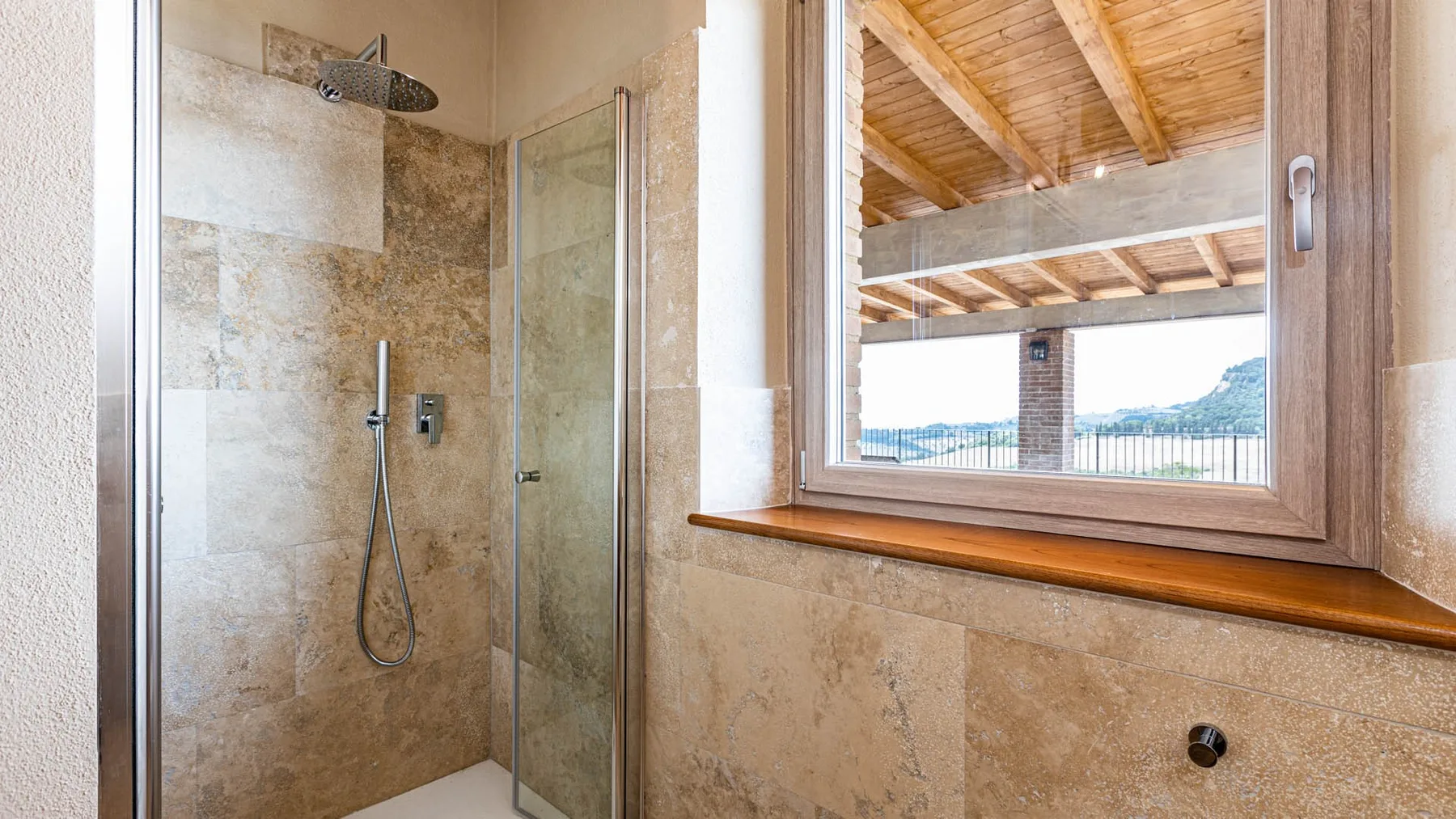 A tiled bathroom with a glass shower beside a window overlooking a covered terrace.