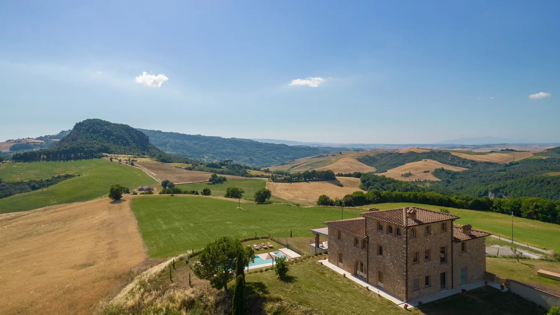A stone house sits beside a small pool amid rolling fields and hills.