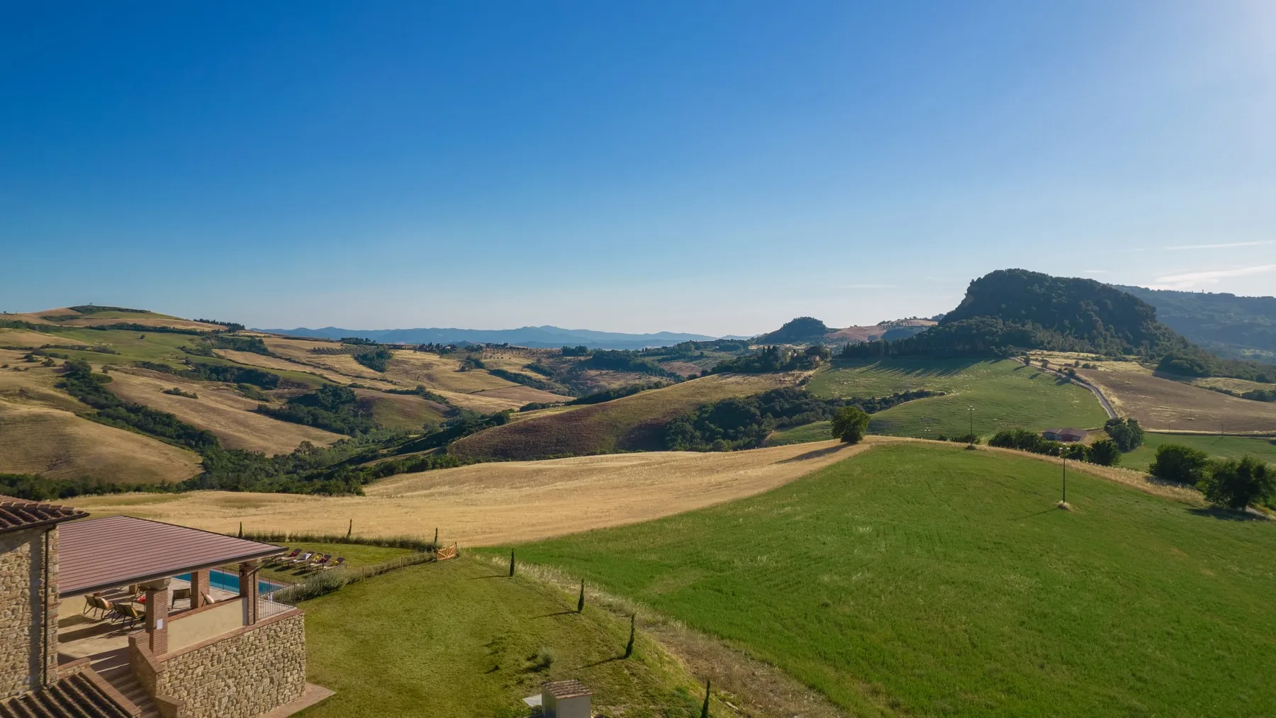 Rolling countryside surrounds green fields and a small covered patio beneath a clear sky.