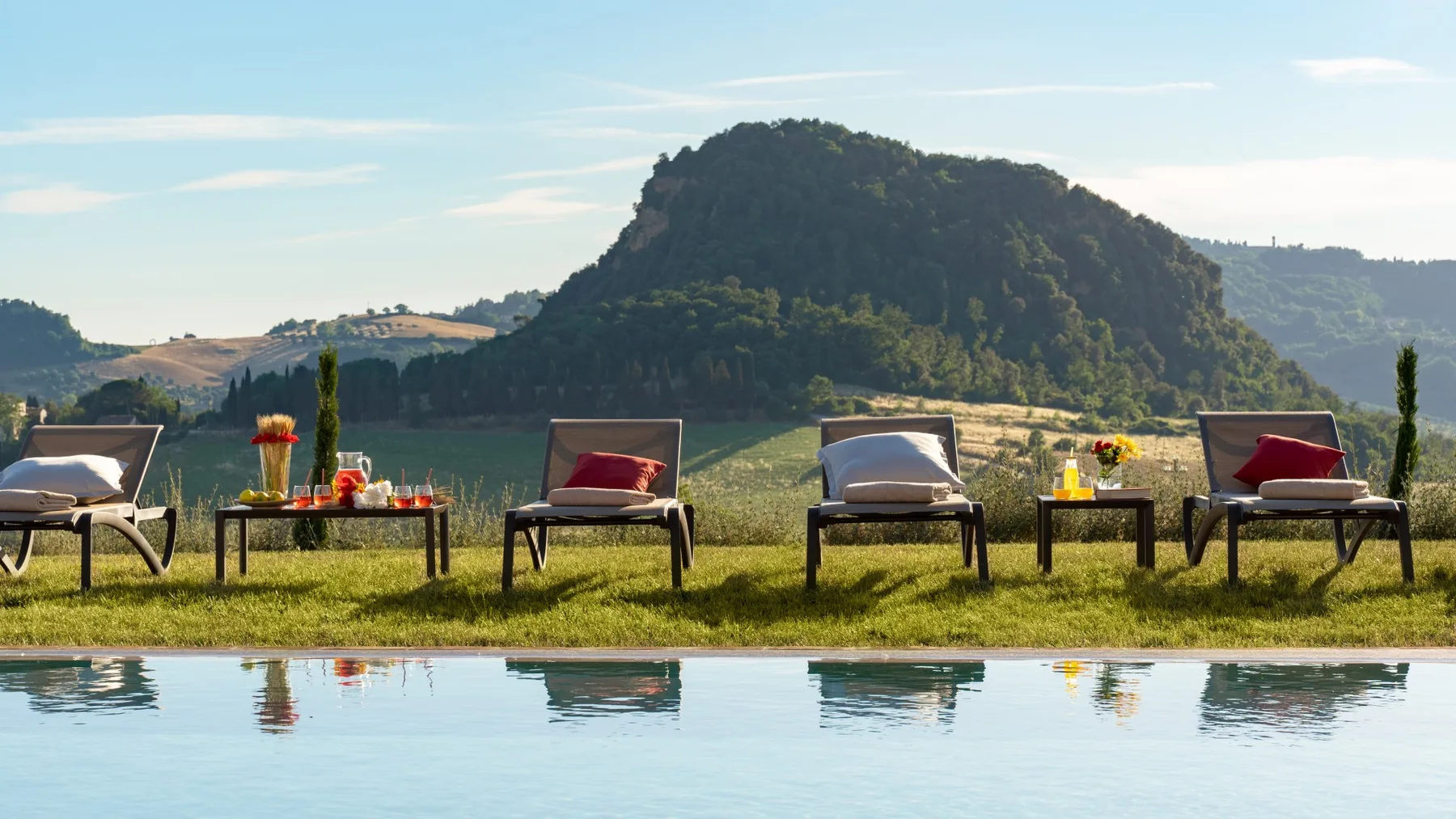 Outdoor seating beside a swimming pool overlooks a green, tree-covered hill.