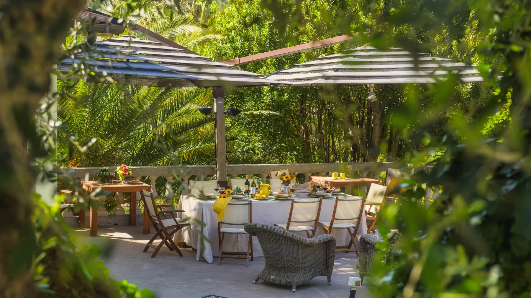 Una zona de comedor al aire libre está bajo toldos a rayas, rodeada de vegetación.