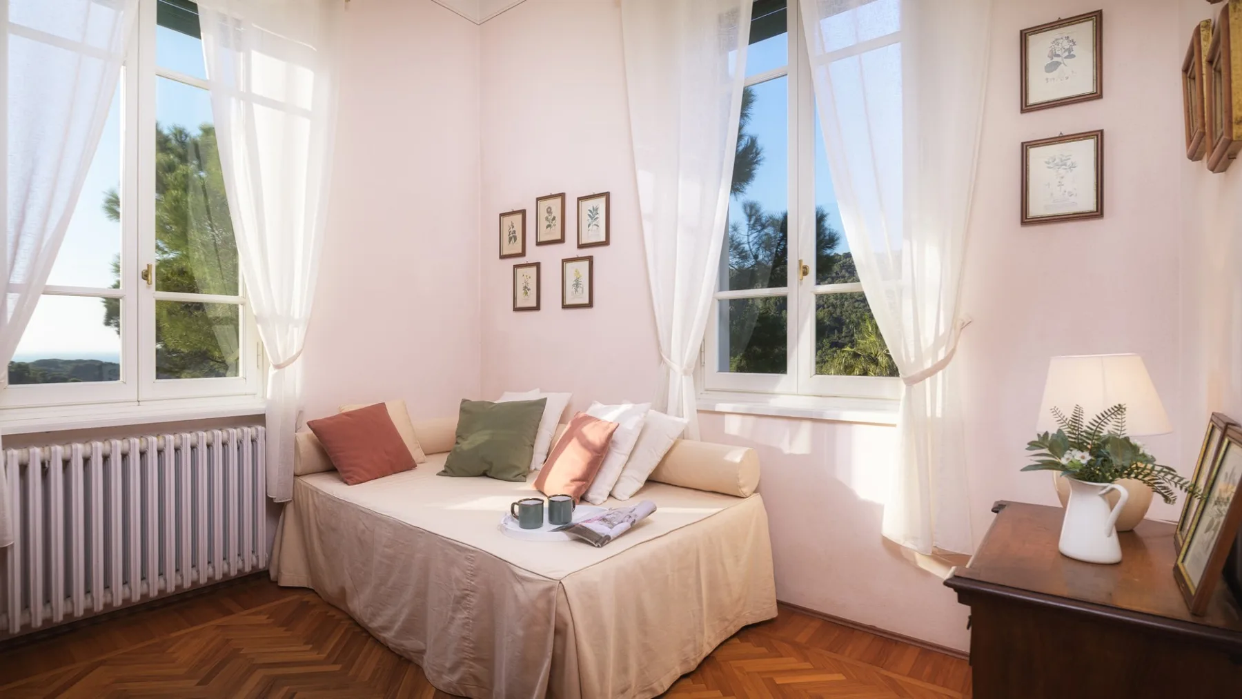 Un dormitorio luminoso con una cama bajo grandes ventanas y cortinas transparentes.