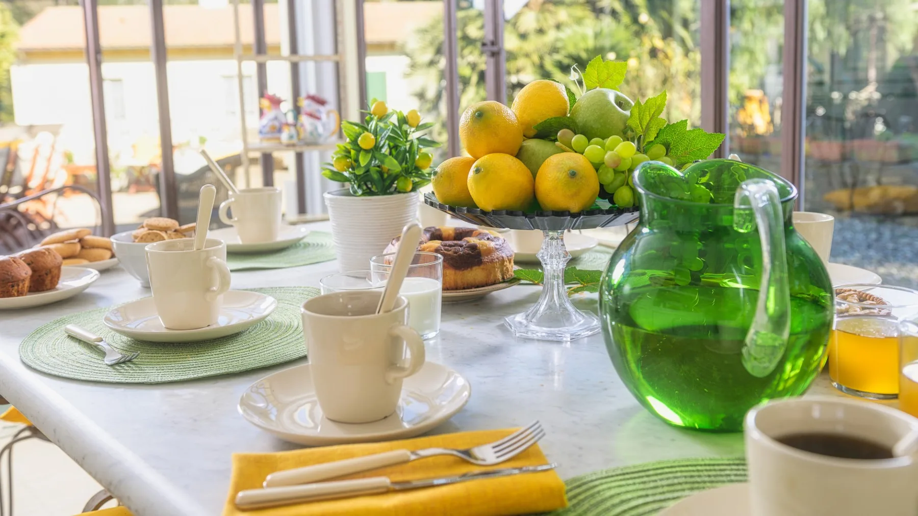 Una mesa de desayuno con tazas, pasteles, fruta y una jarra de vidrio verde.