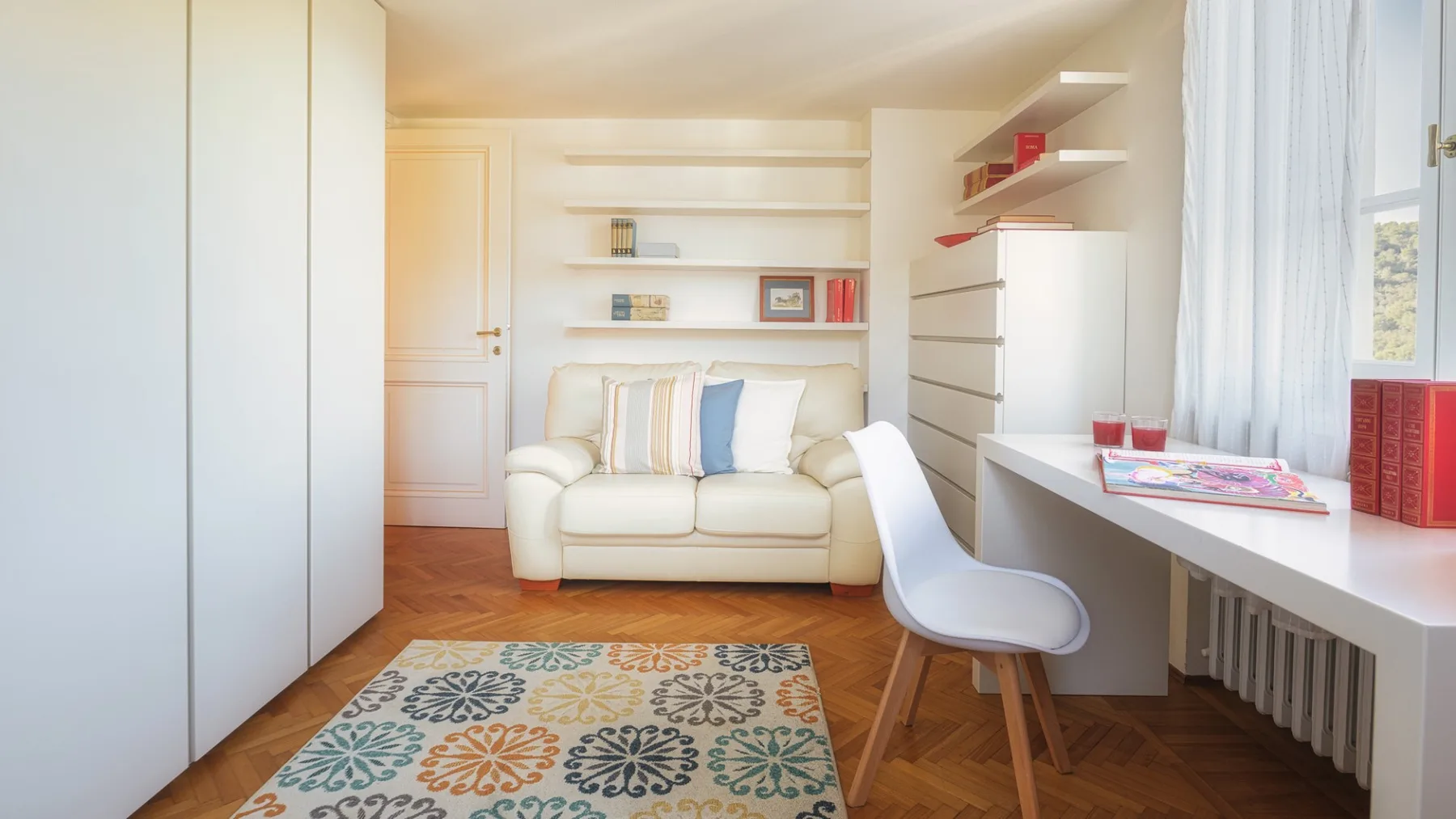 Una habitación luminosa con un sofá crema, estantes en la pared y un escritorio junto a la ventana.