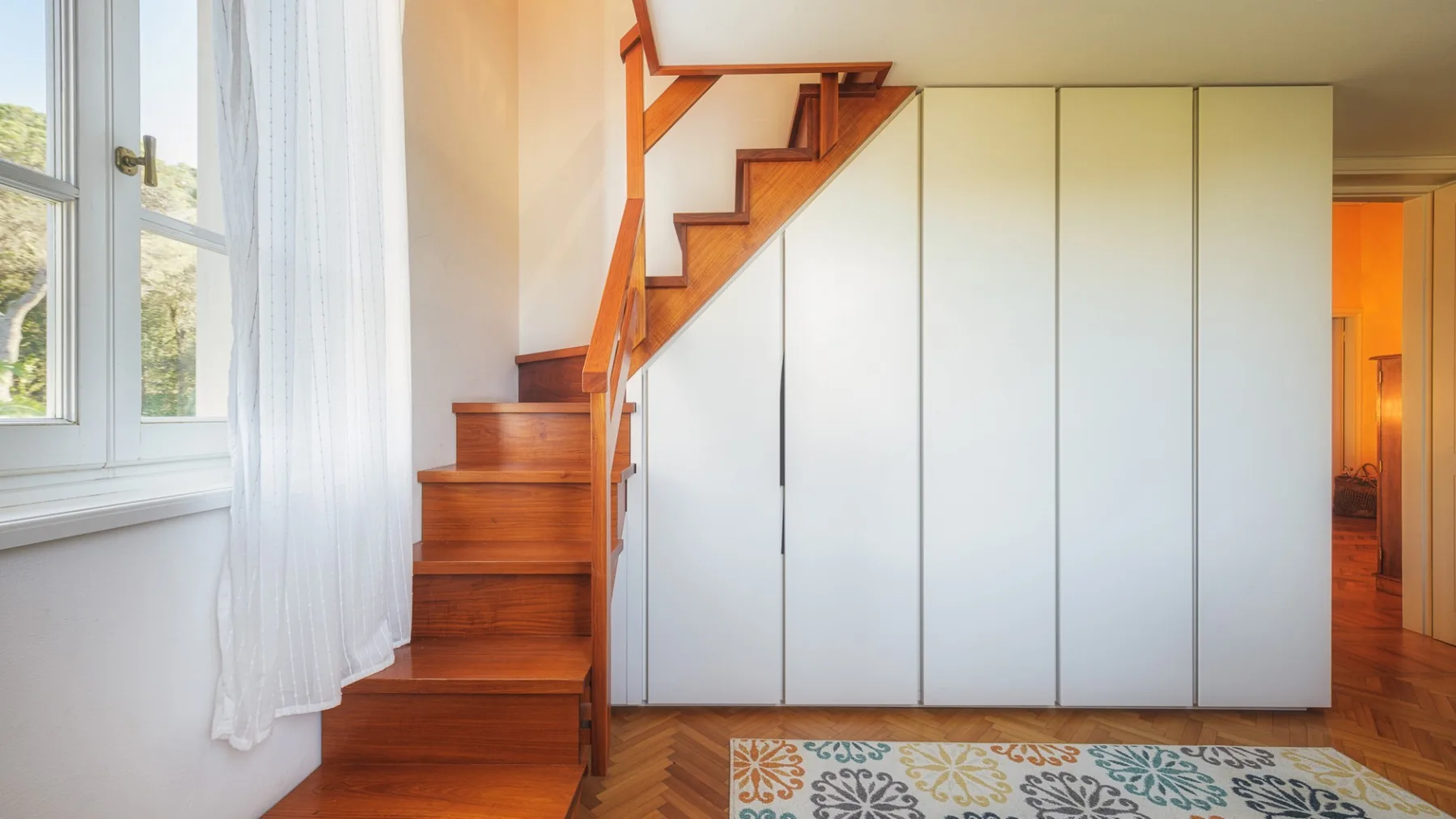 Un interior luminoso muestra una escalera de madera junto a armarios blancos altos.