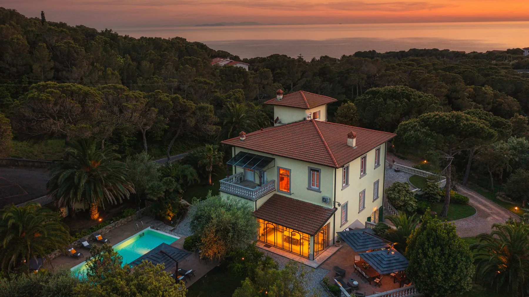 Una casa grande entre árboles da a una piscina y la costa al atardecer.