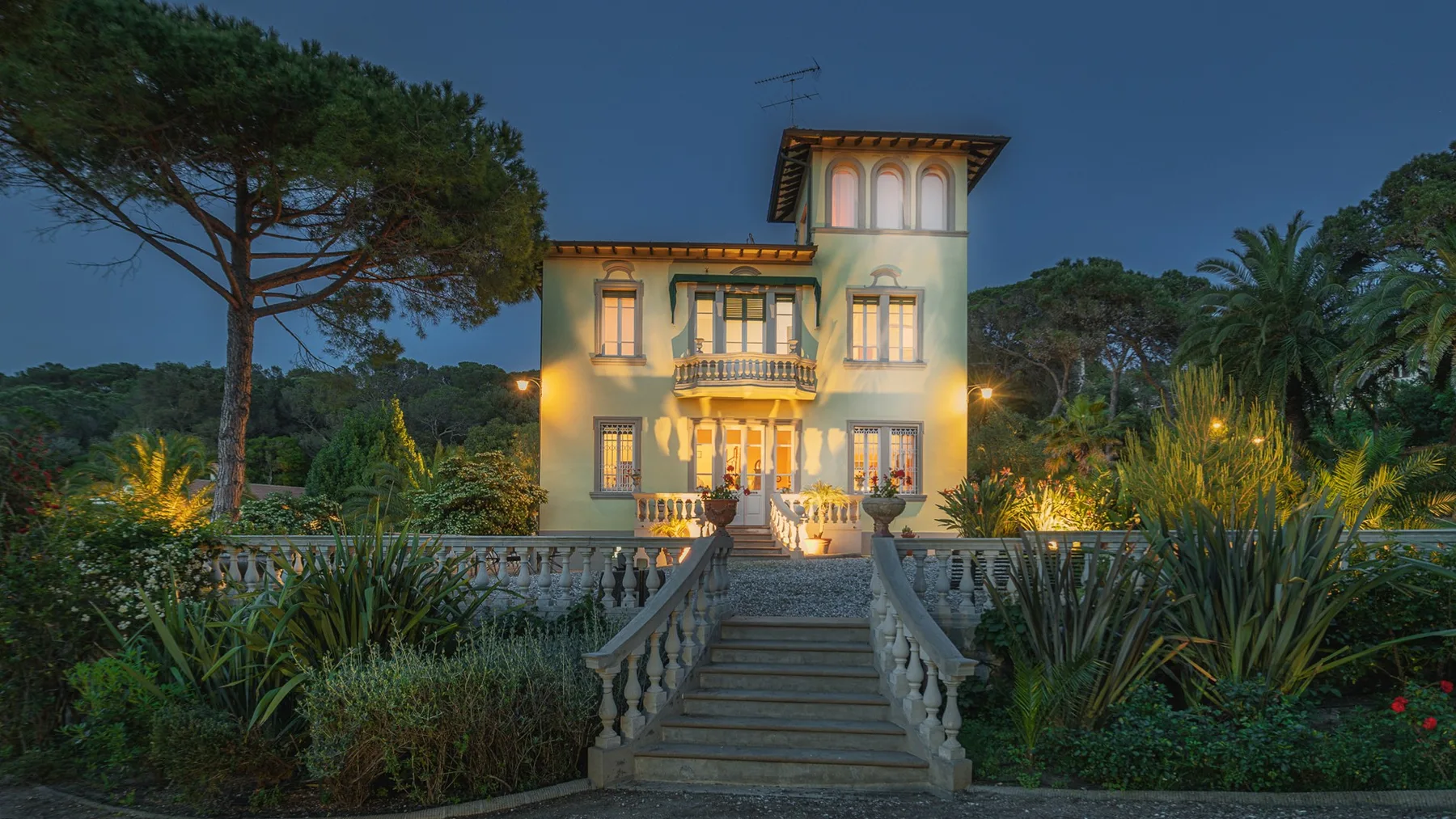 Una villa iluminada se alza tras una escalera de piedra, rodeada de jardines al anochecer.