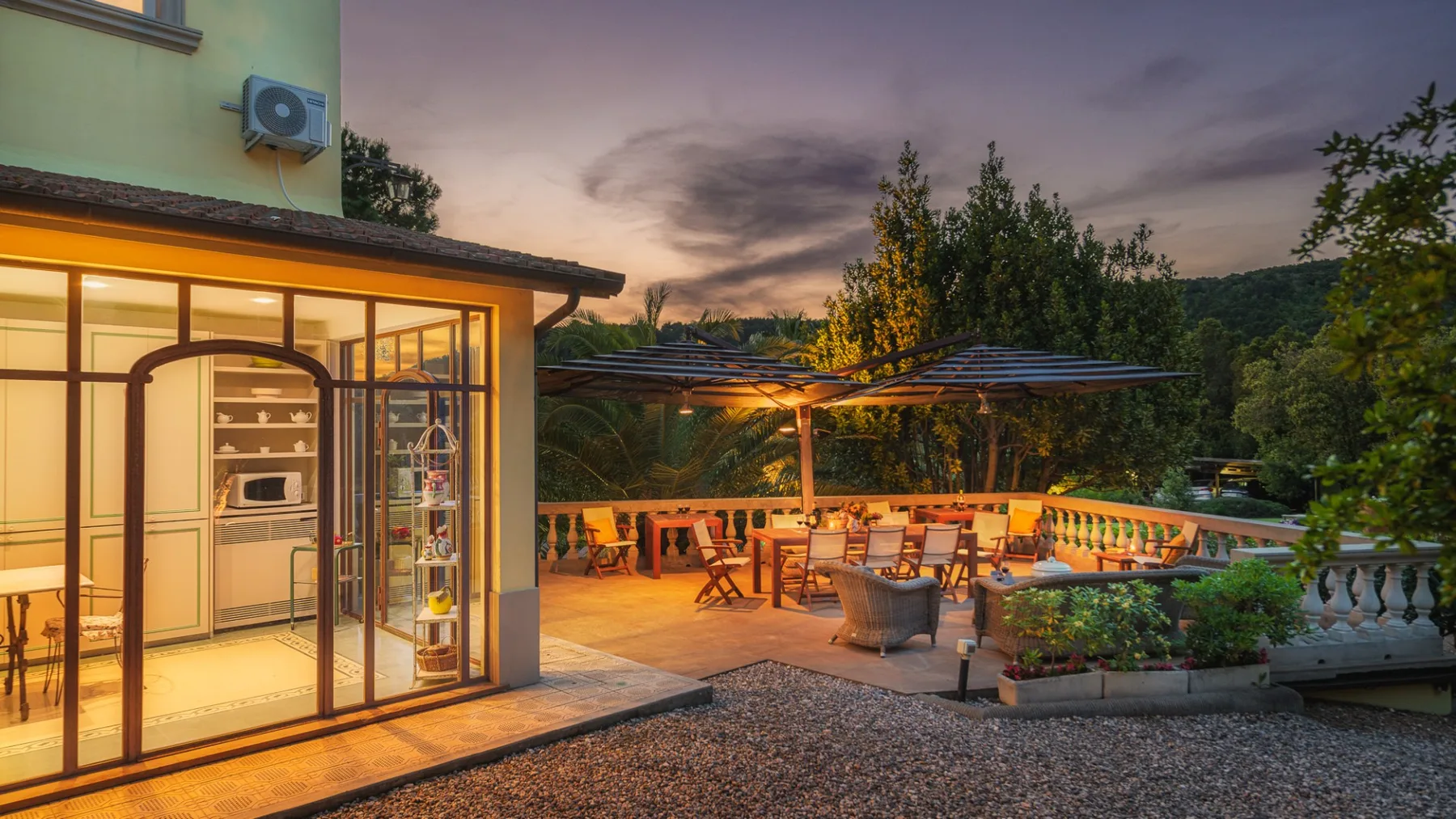 Una zona de comedor en un patio iluminado junto a una cocina acristalada al anochecer.