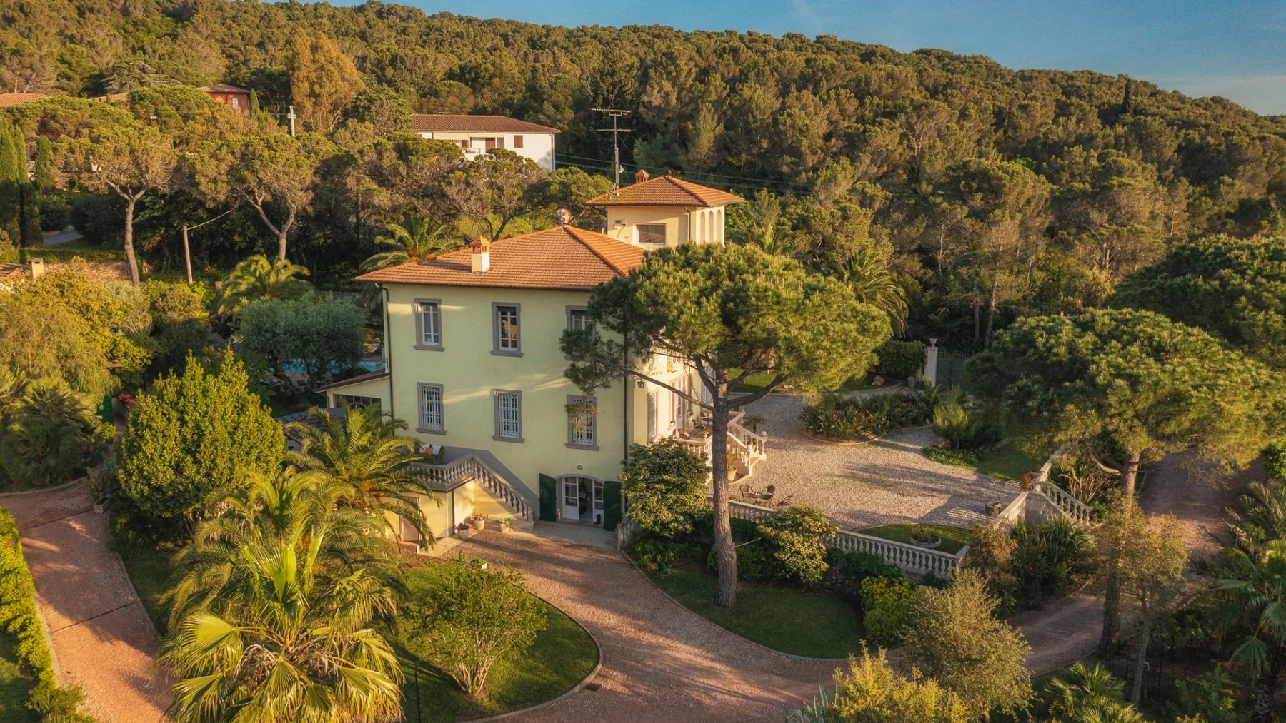 Una casa clara con techo de terracota está rodeada de árboles y jardines.