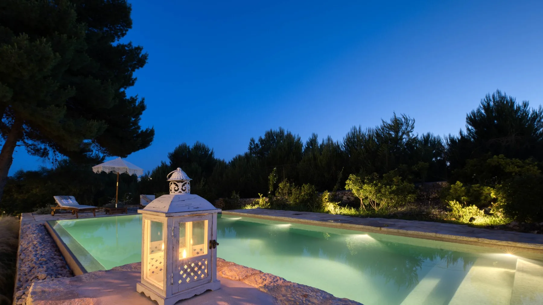 Una piscina al aire libre brilla al anochecer junto a un farol decorativo.