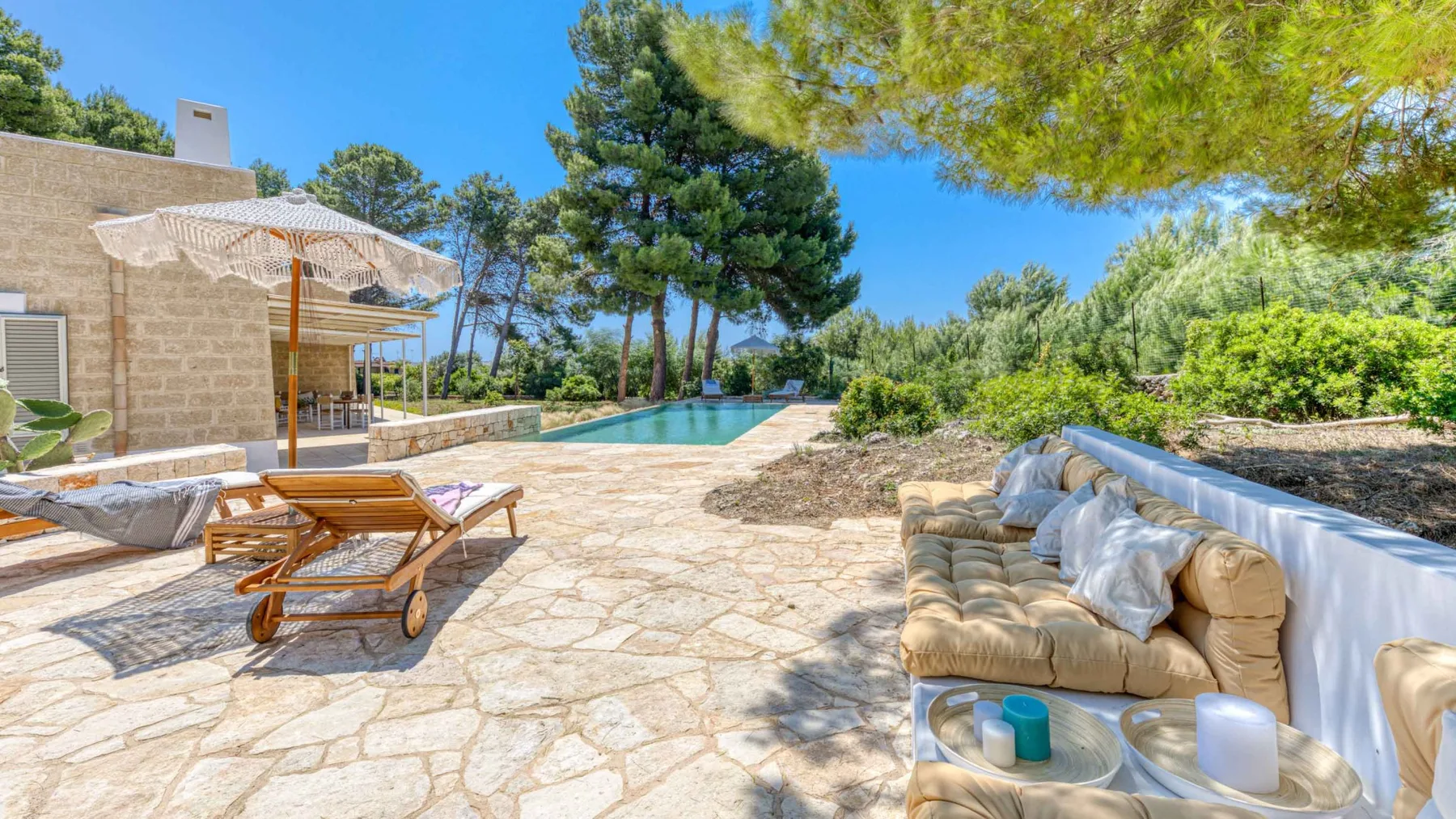 Un patio soleado de piedra con asientos al aire libre, una piscina y árboles alrededor.