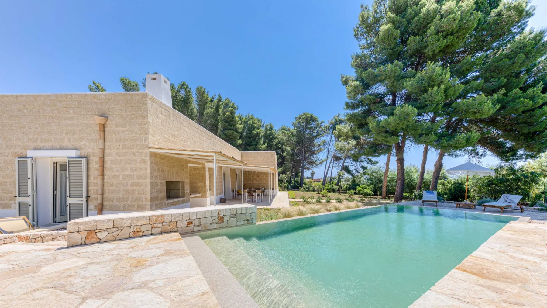 Una piscina junto a una casa de piedra clara y árboles altos.
