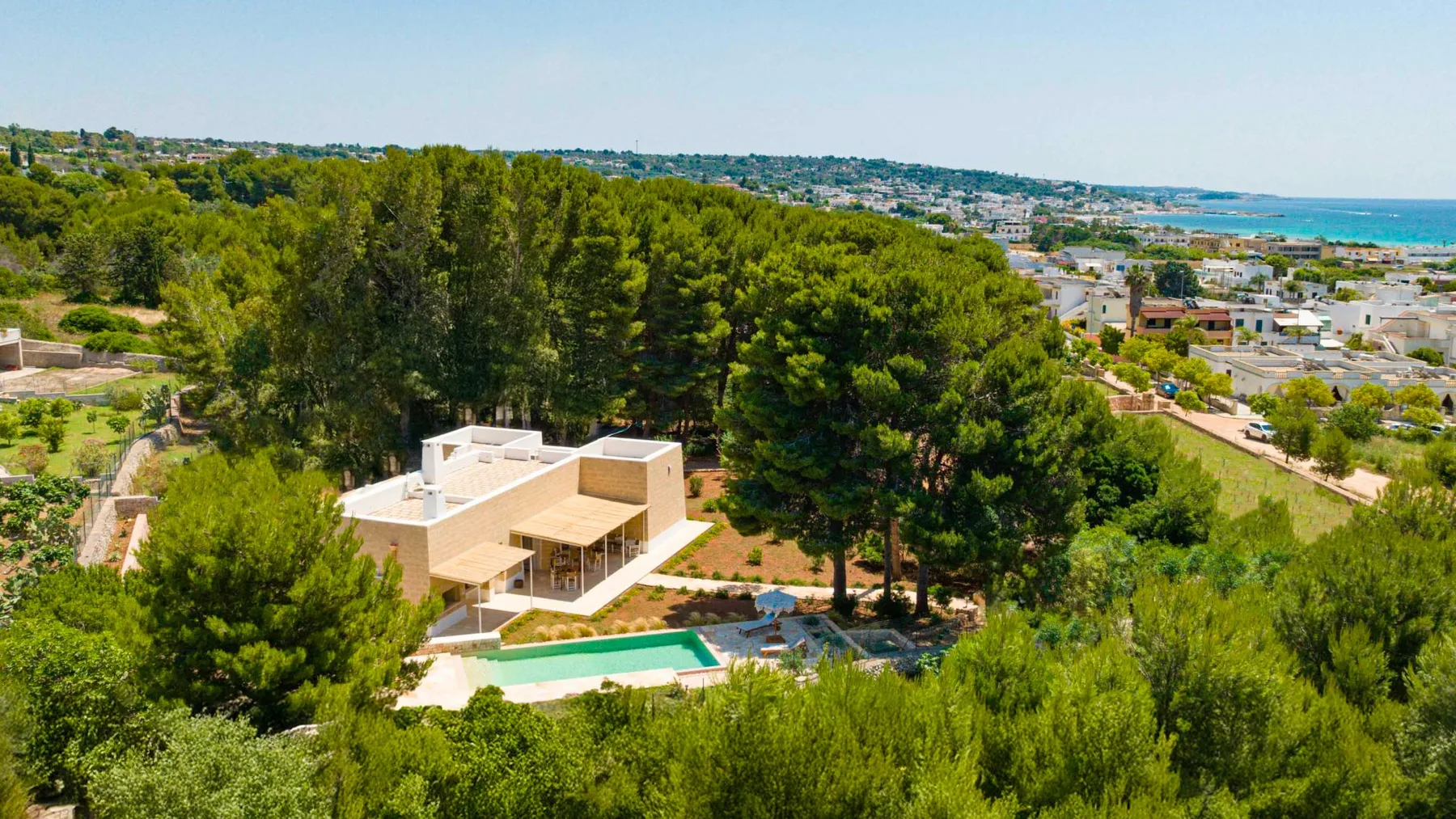 Una casa moderna y una piscina se encuentran entre árboles, con vistas a un pueblo costero.