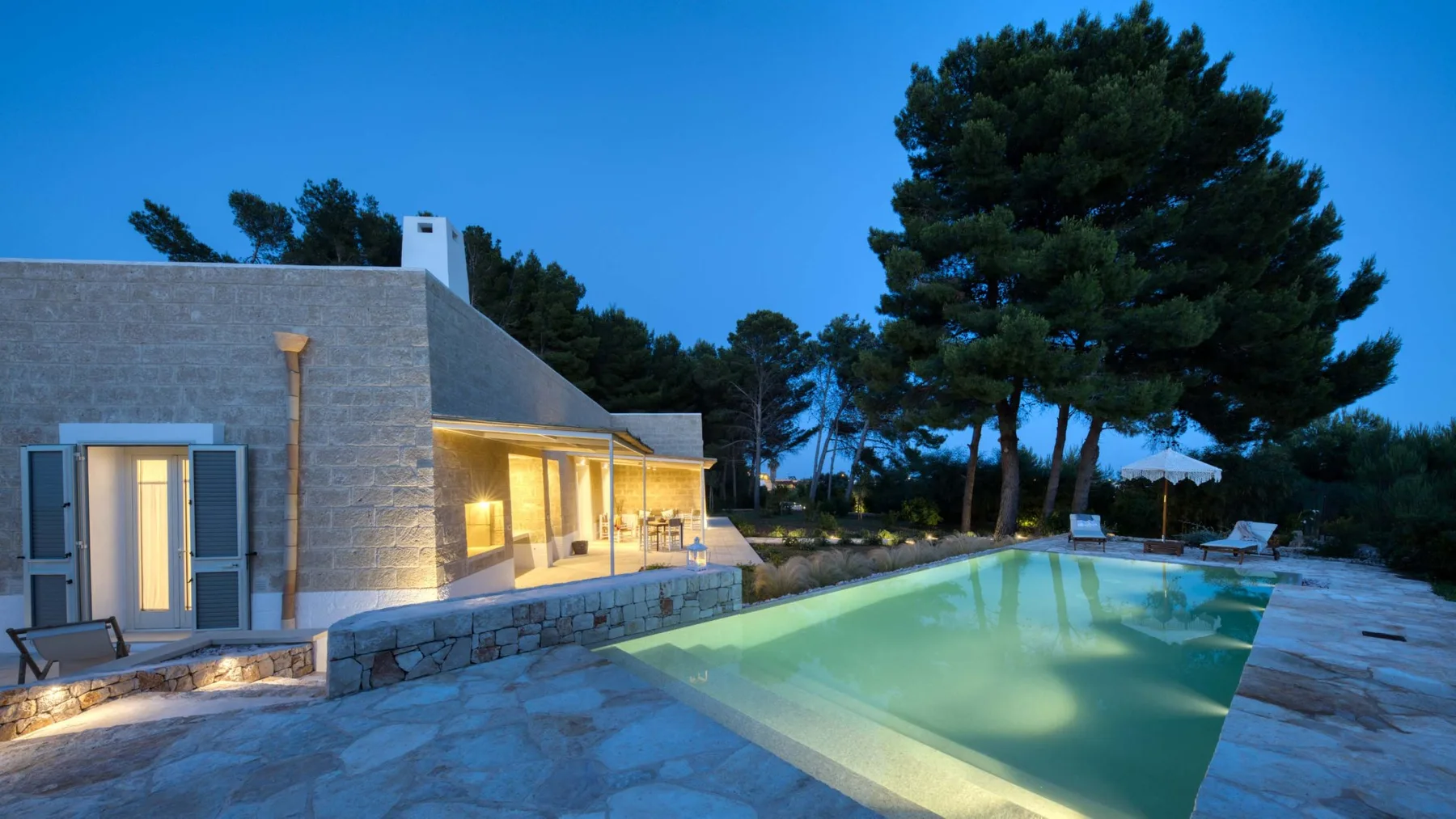 Una casa moderna de piedra y una piscina iluminada aparecen al anochecer.