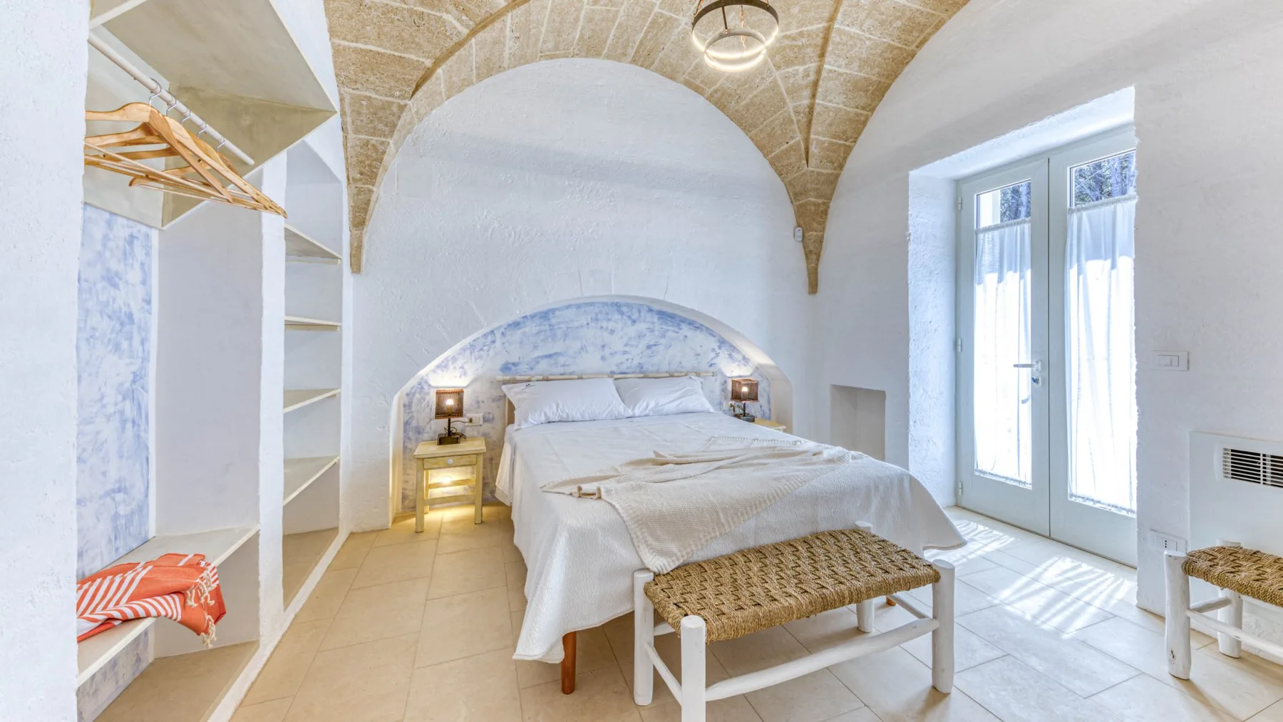 Un dormitorio luminoso con una cama, un banco tejido y ventanas altas.