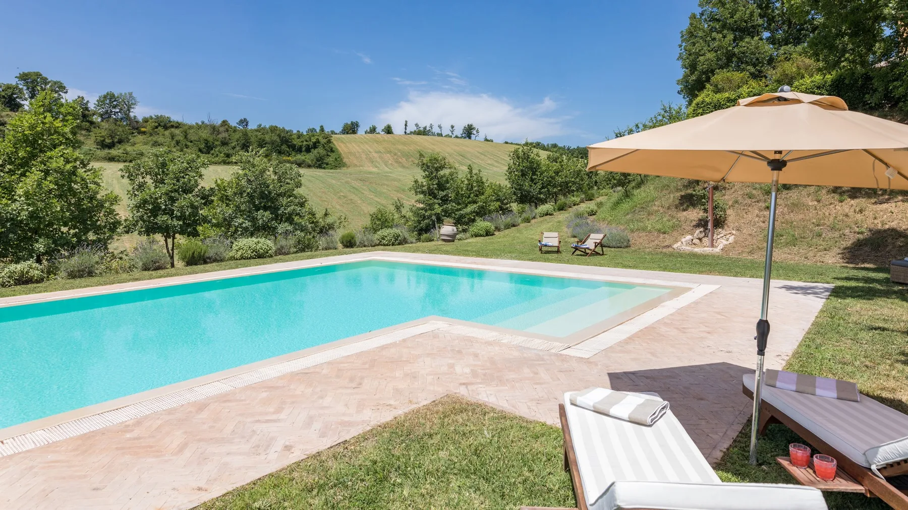 An outdoor swimming pool sits beside a paved deck and hillside greenery.