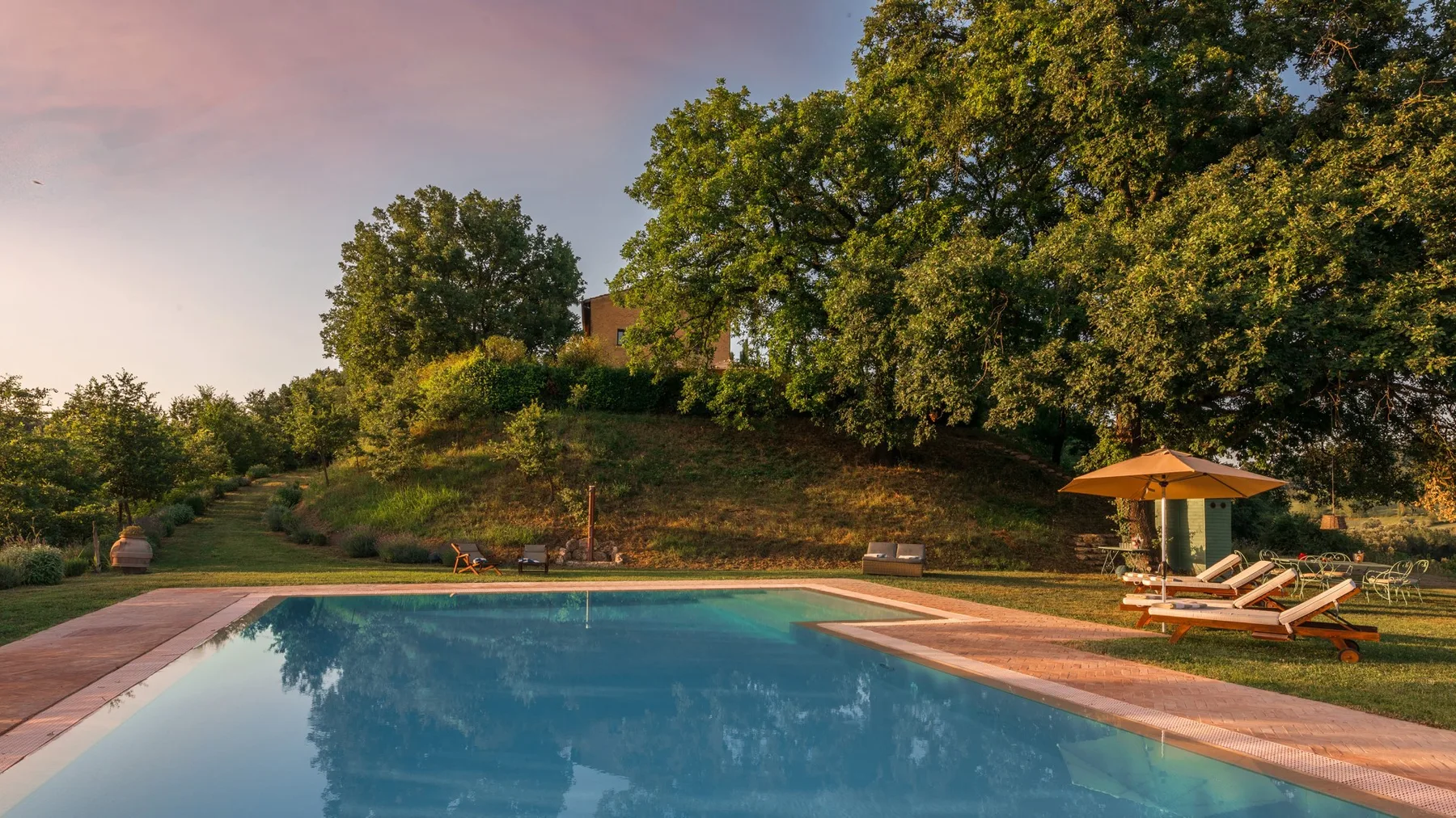 An outdoor swimming pool sits beside a grassy slope and leafy trees.