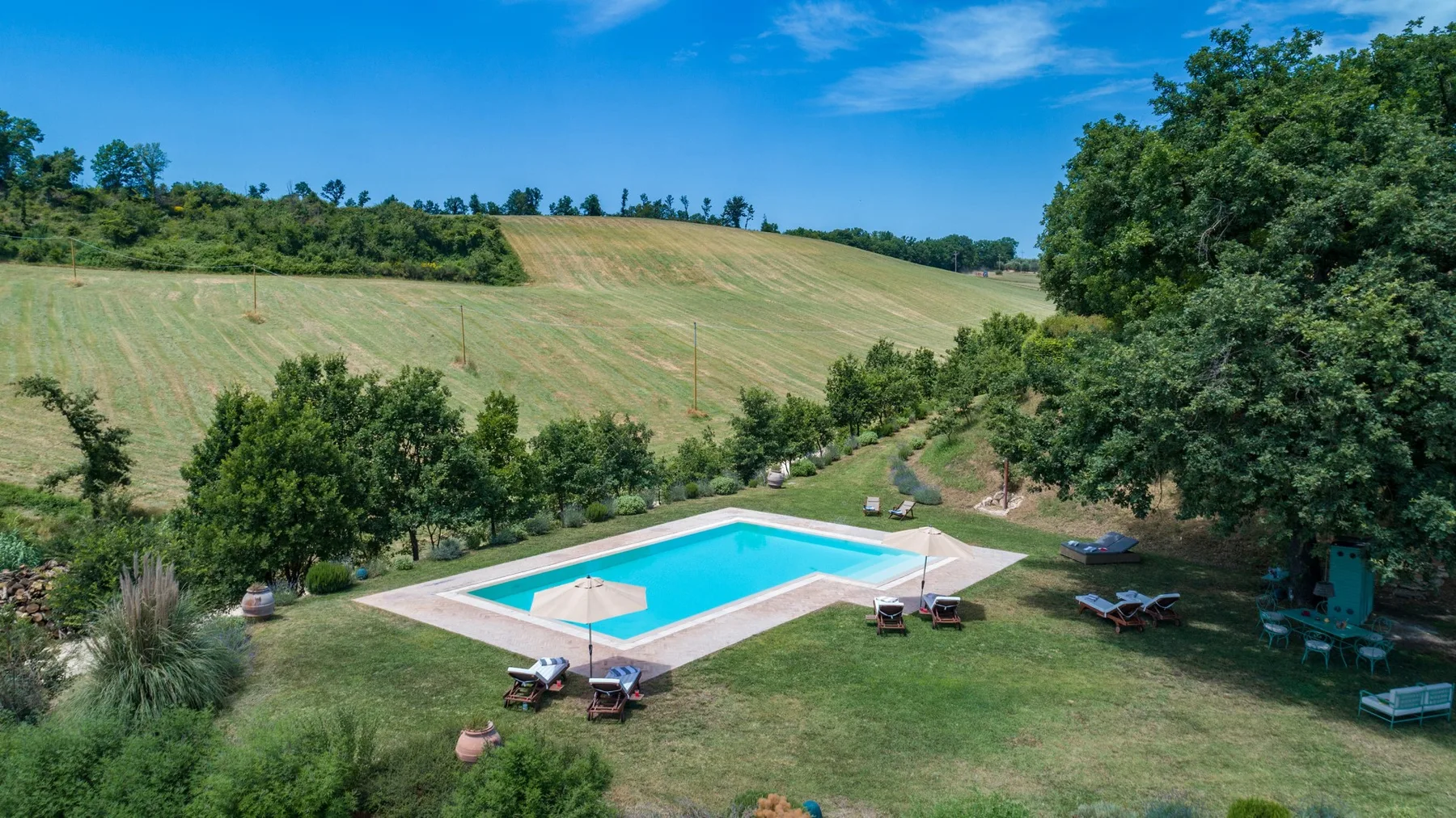 An outdoor swimming pool sits in a grassy yard bordered by trees and open fields.