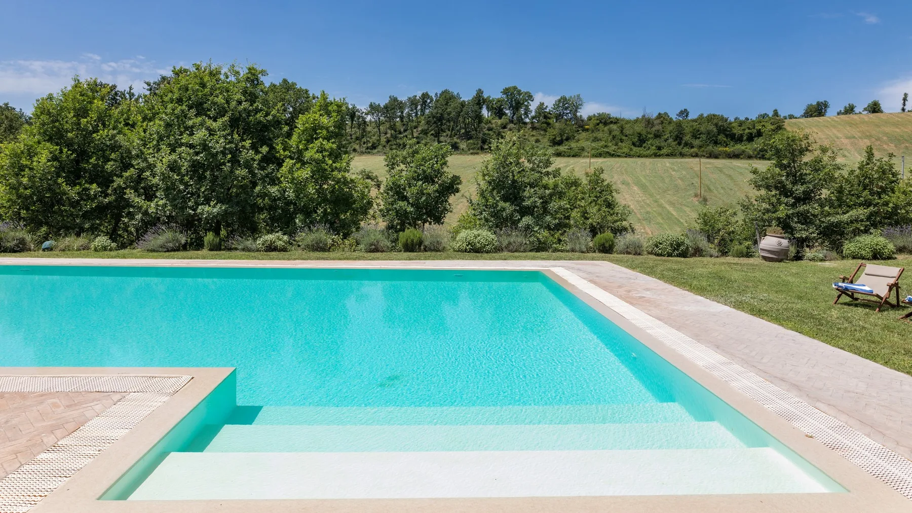 An outdoor swimming pool bordered by a paved deck and green countryside.