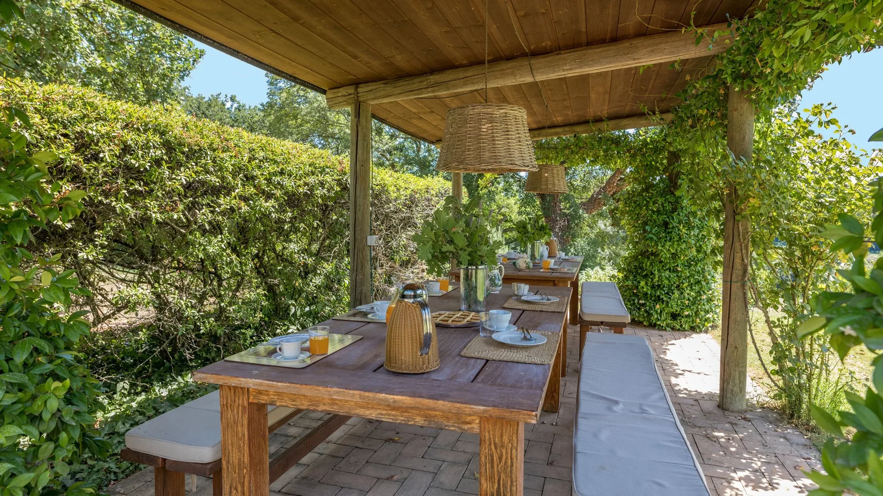 A covered outdoor dining area with a long wooden table surrounded by greenery.
