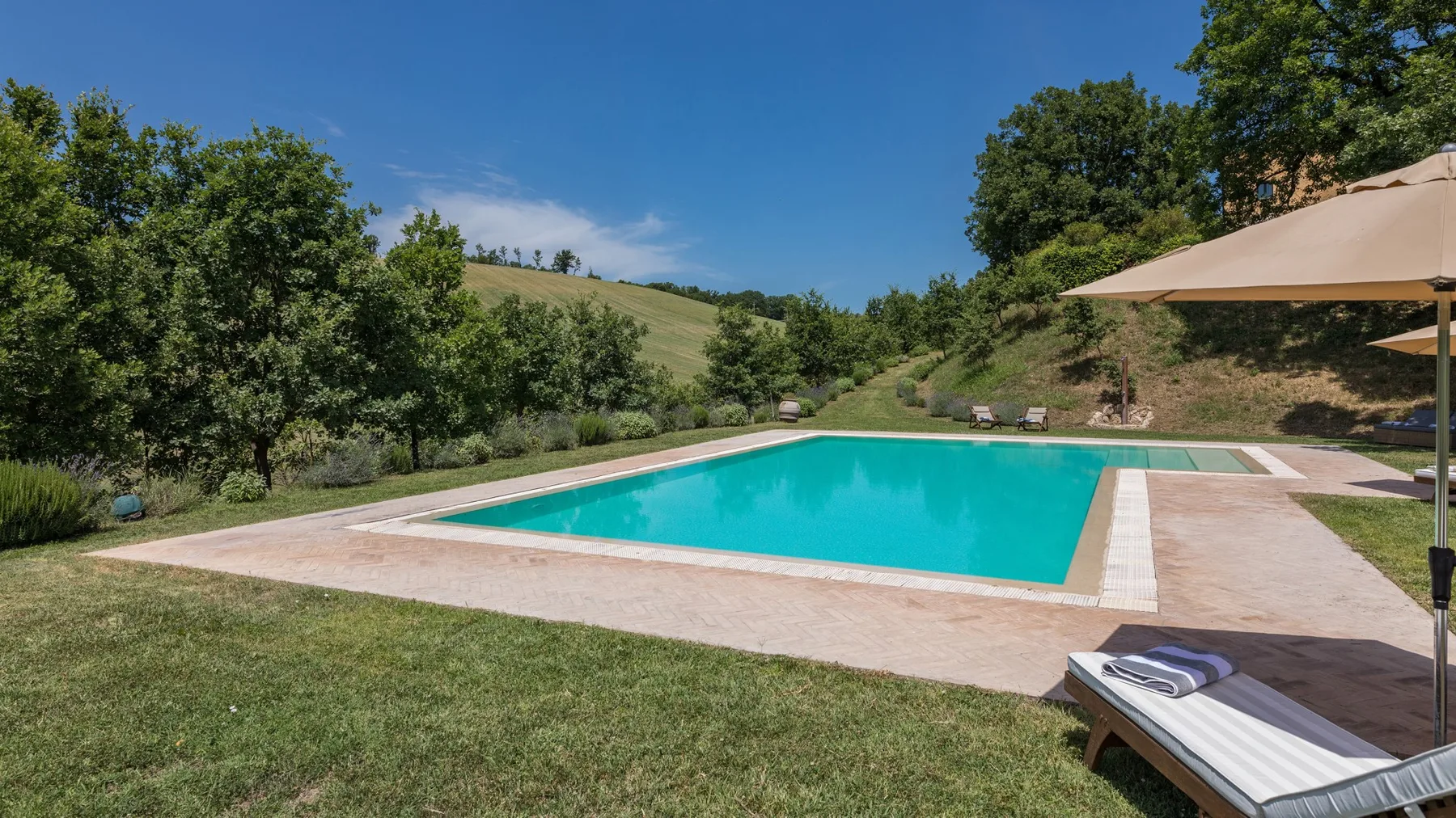 An outdoor swimming pool sits beside a grassy hillside and trees.