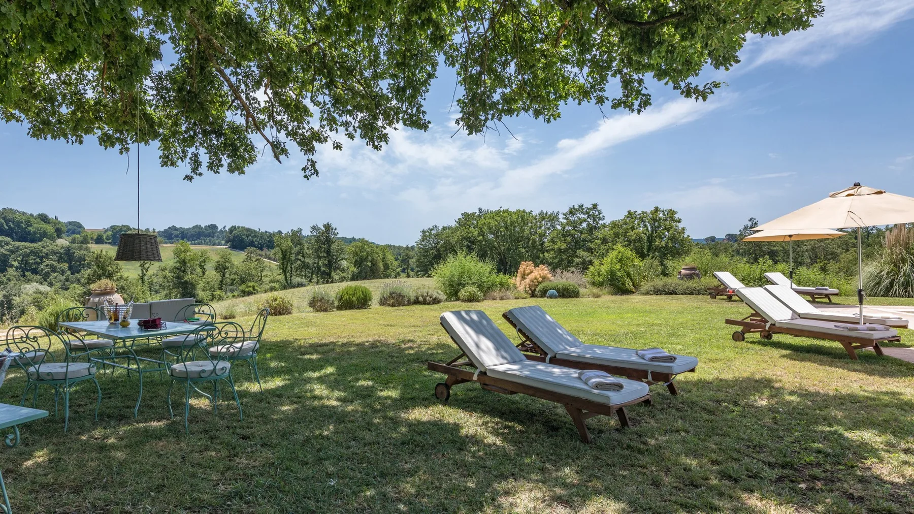 An outdoor garden seating area overlooks open fields, with lounge chairs beneath a leafy tree.