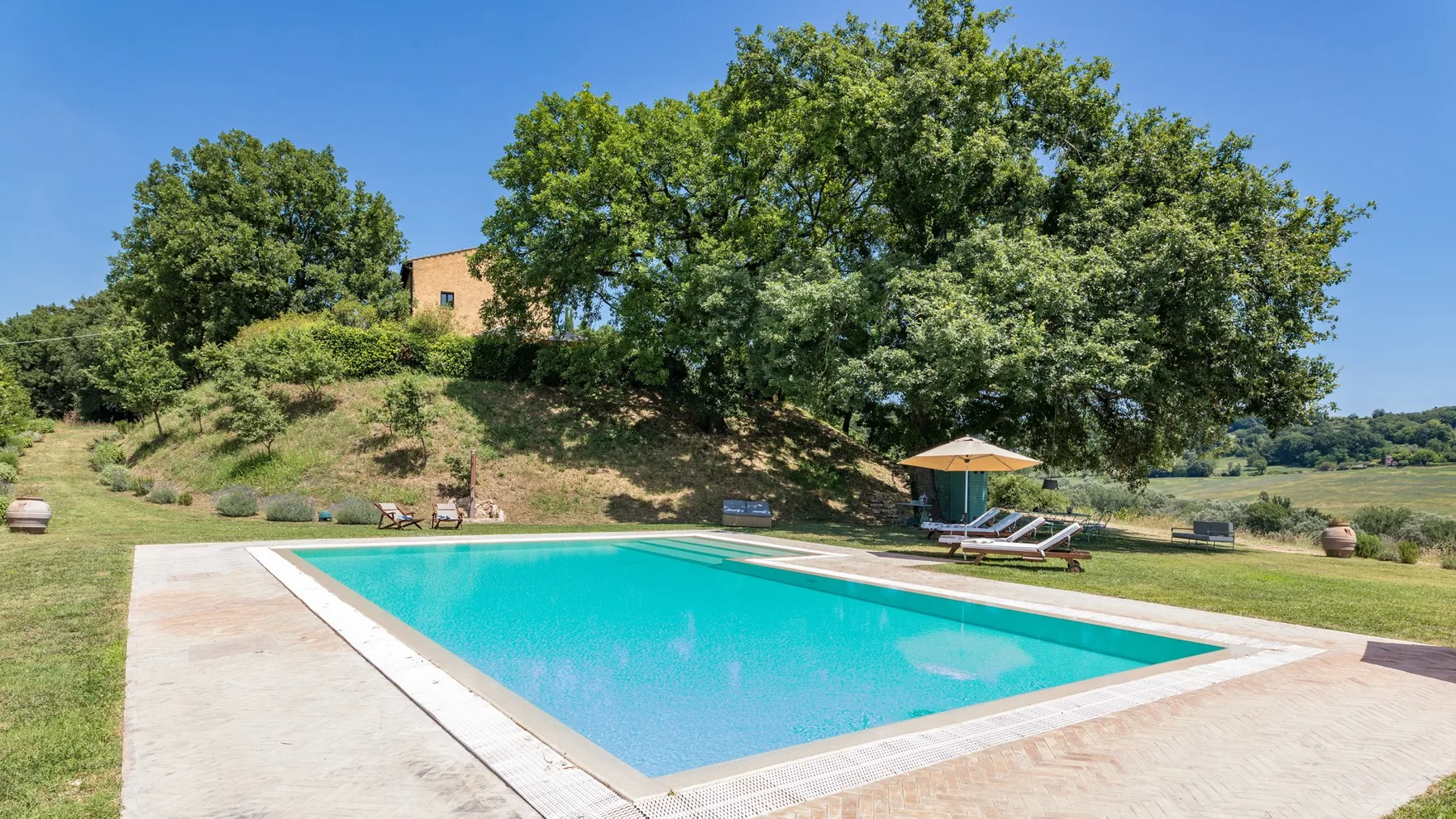 An outdoor swimming pool sits beside a grassy, tree-lined slope.