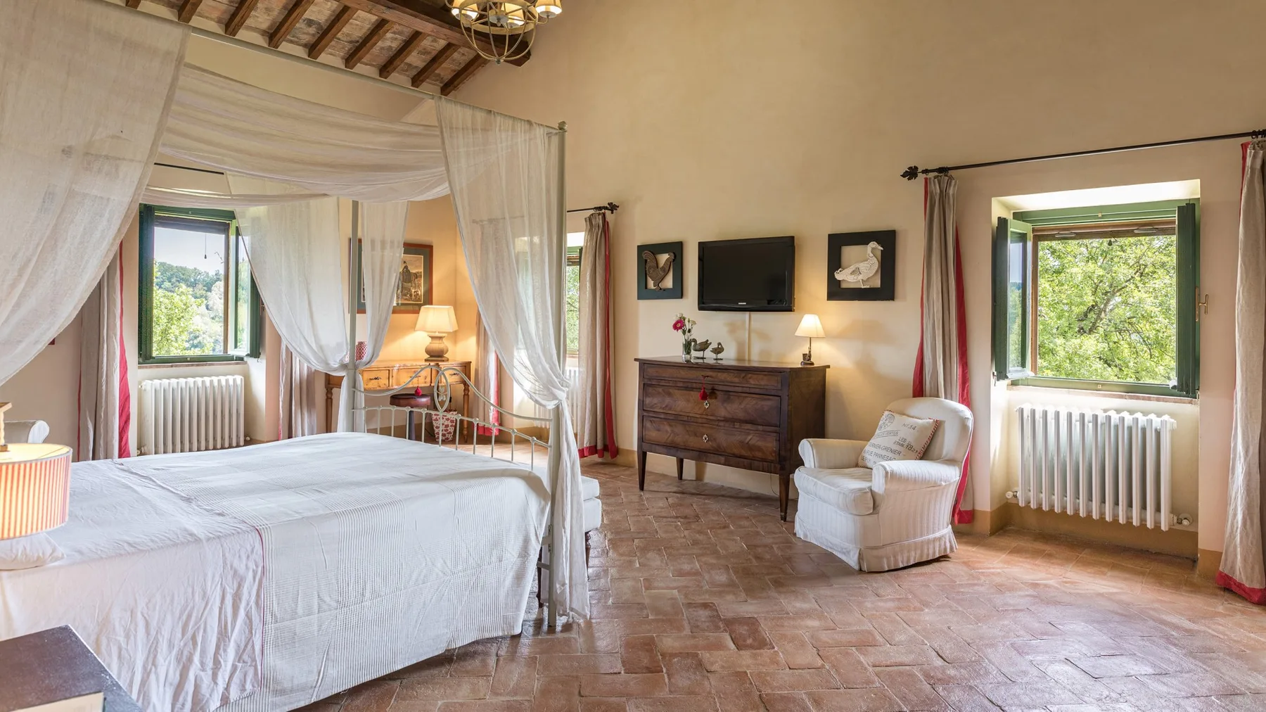 A bedroom with a large draped bed, a dresser, and windows overlooking greenery.