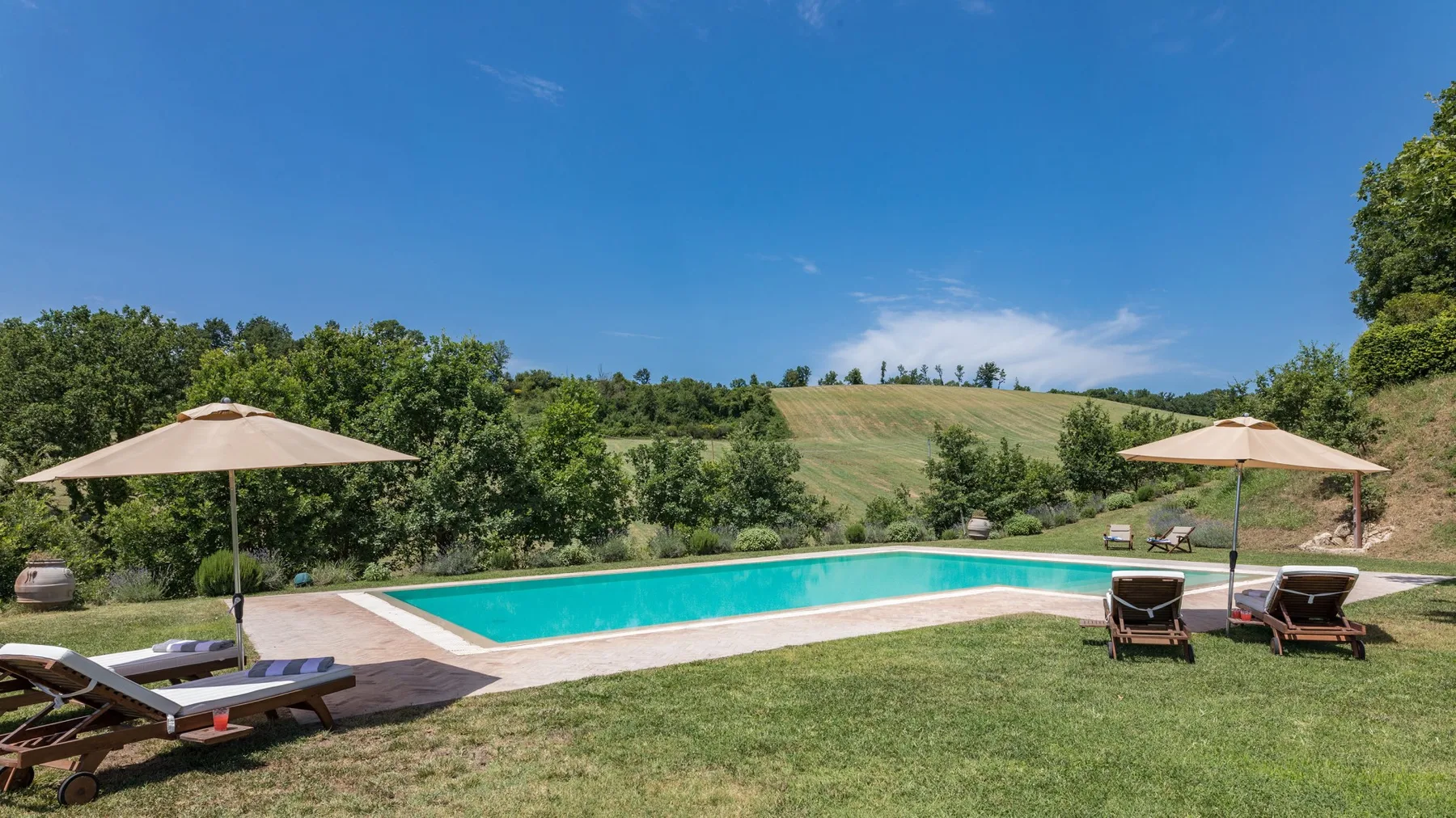 An outdoor swimming pool is surrounded by lawn, lounge seating, and green hills.
