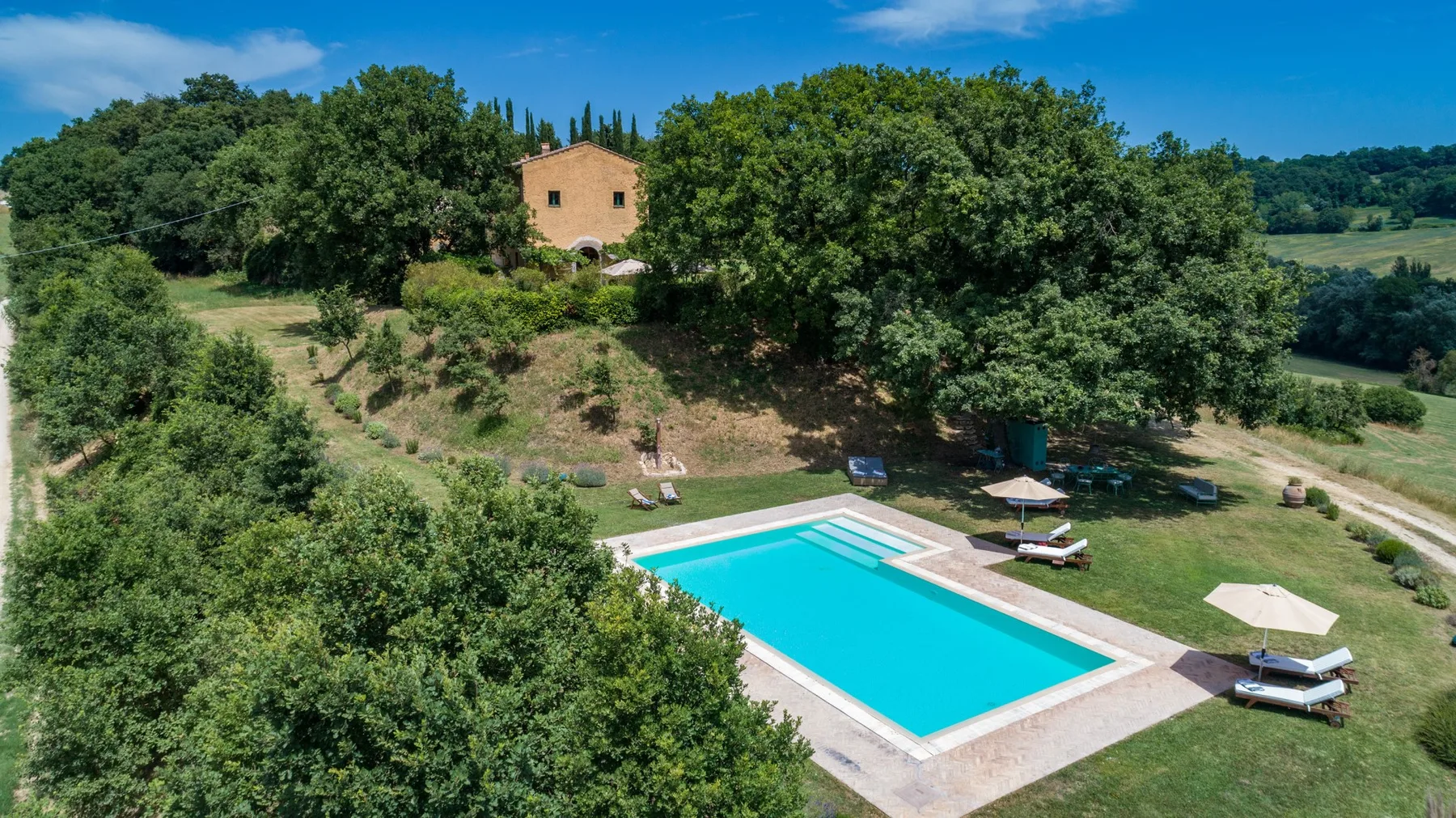 A swimming pool sits beside a grassy yard and tree-covered hillside.