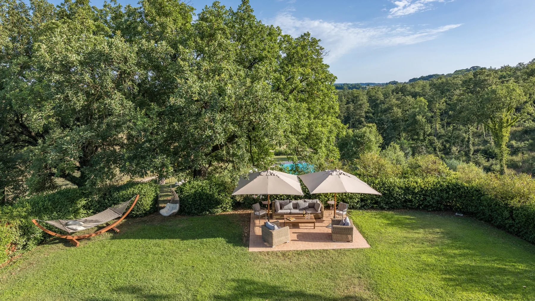 A garden patio with shaded seating overlooks a green lawn and wooded hills.