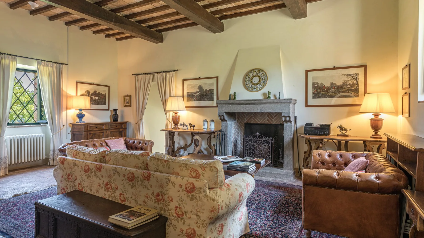 A living room with a fireplace, patterned seating, and exposed wooden ceiling beams.