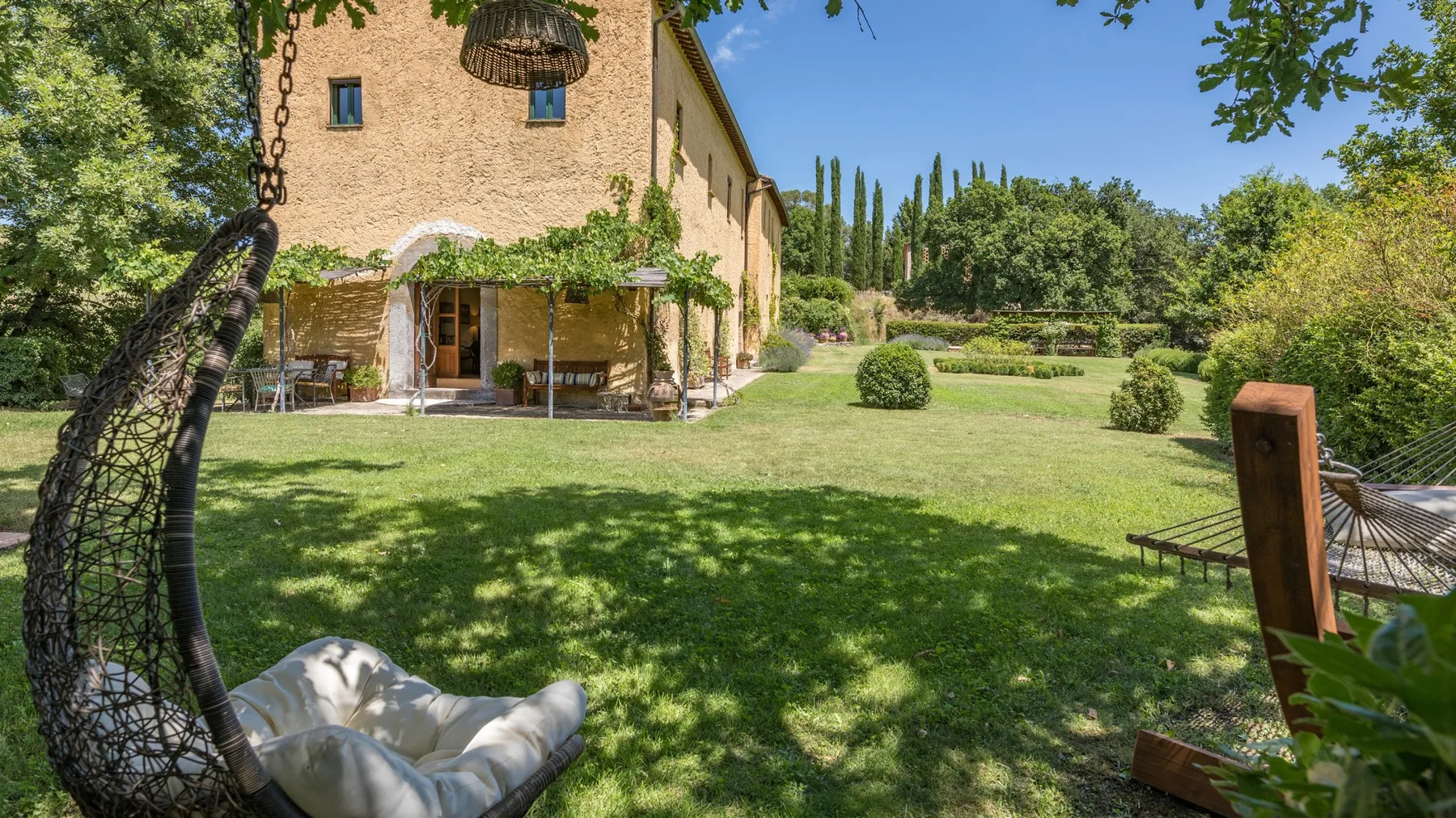 A landscaped garden surrounds a stucco house, with cushioned outdoor seating beneath the trees.