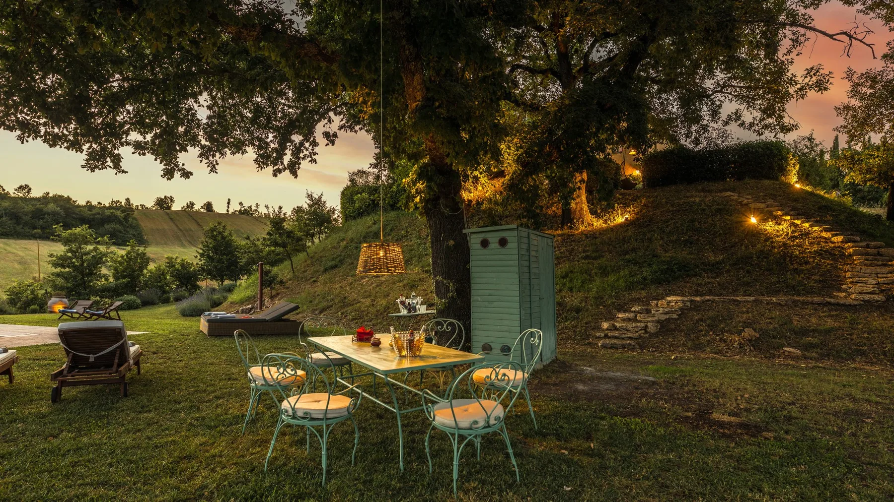 An outdoor dining area sits beneath a large tree, with chairs around a table.