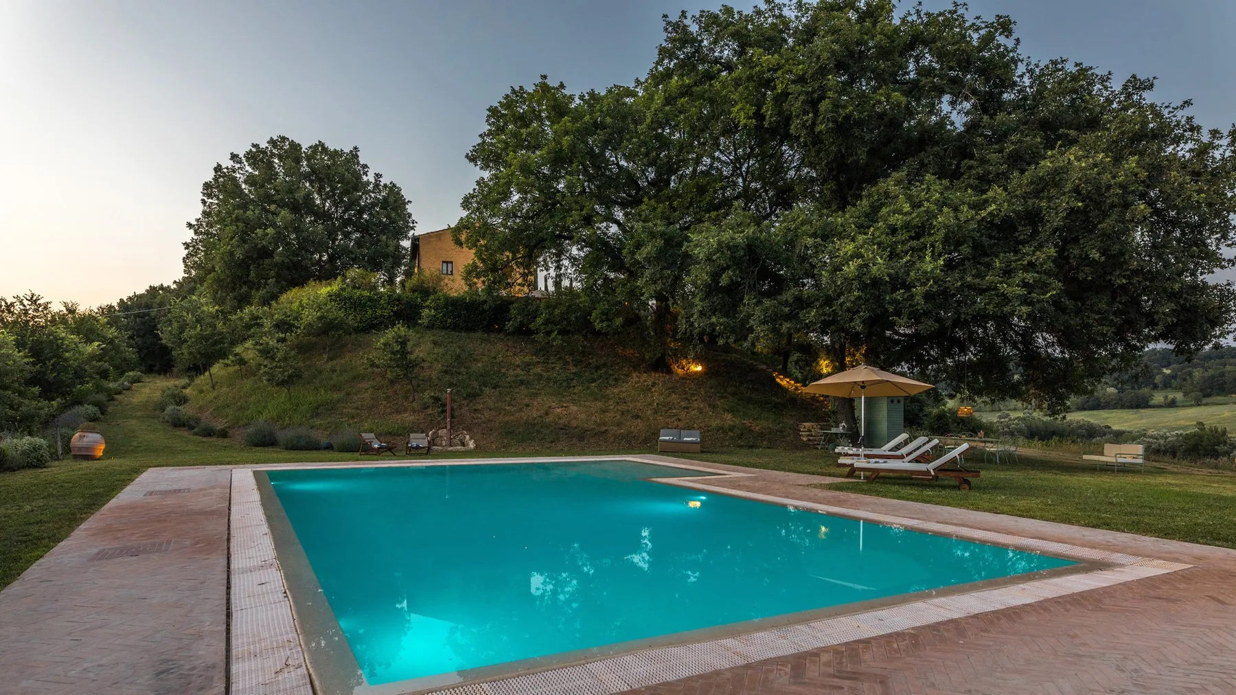 An outdoor swimming pool sits beside a grassy, tree-lined hillside.