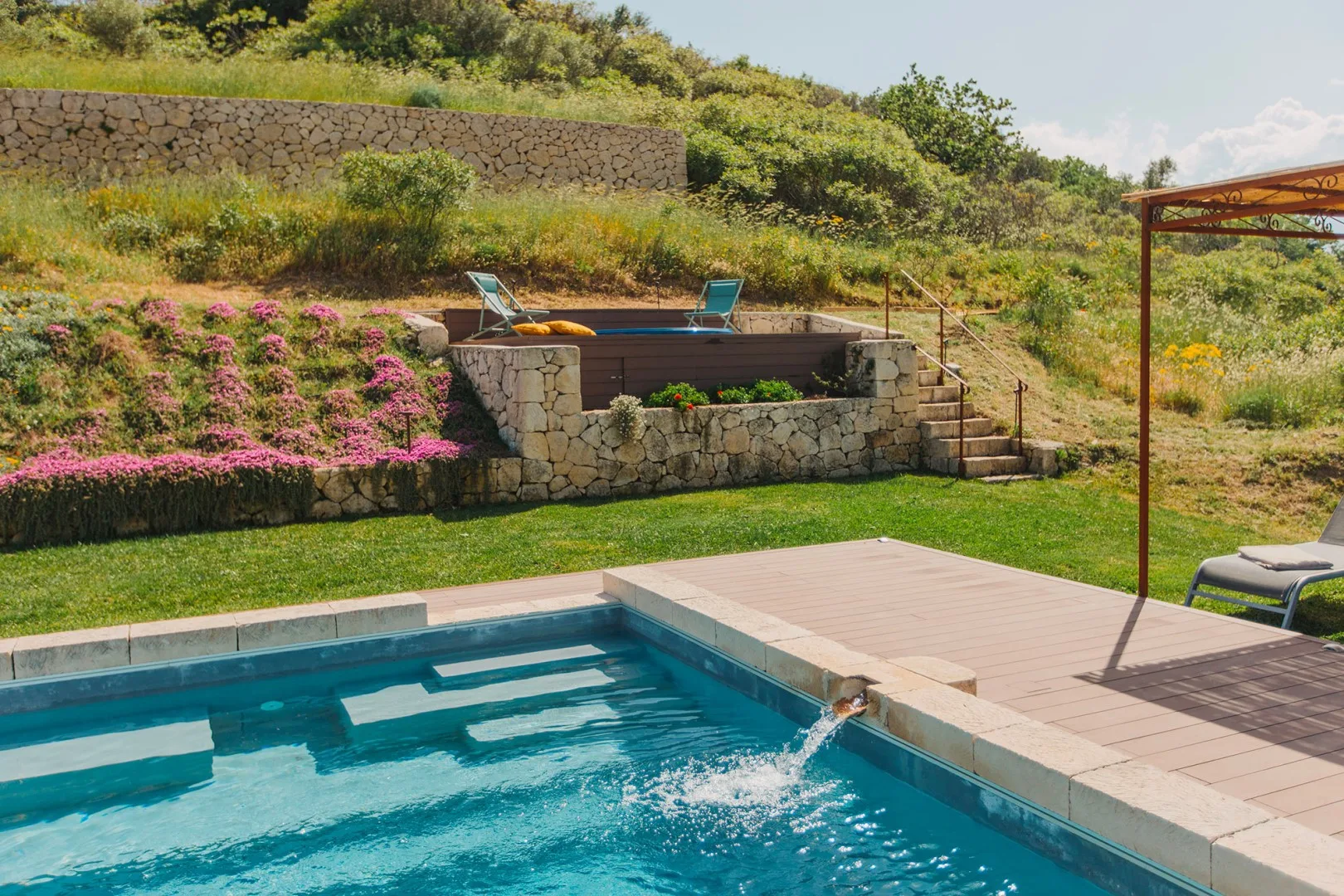 Una piscina exterior está junto a un jacuzzi elevado y una ladera ajardinada.